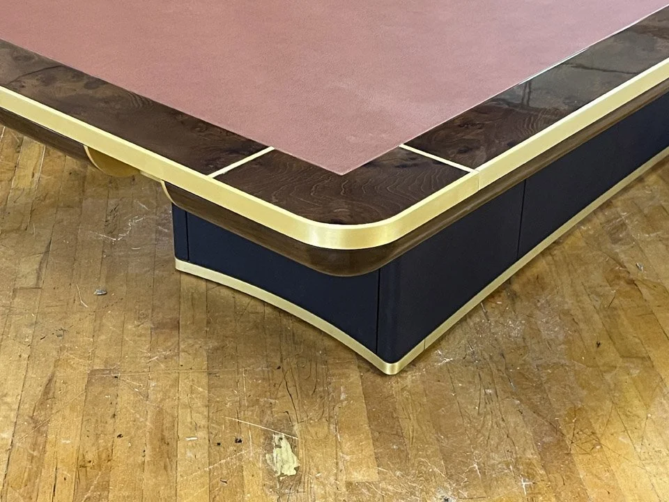 Close-up of a corner of a table with a pink top, dark wood border, and gold trim, on a wooden floor.