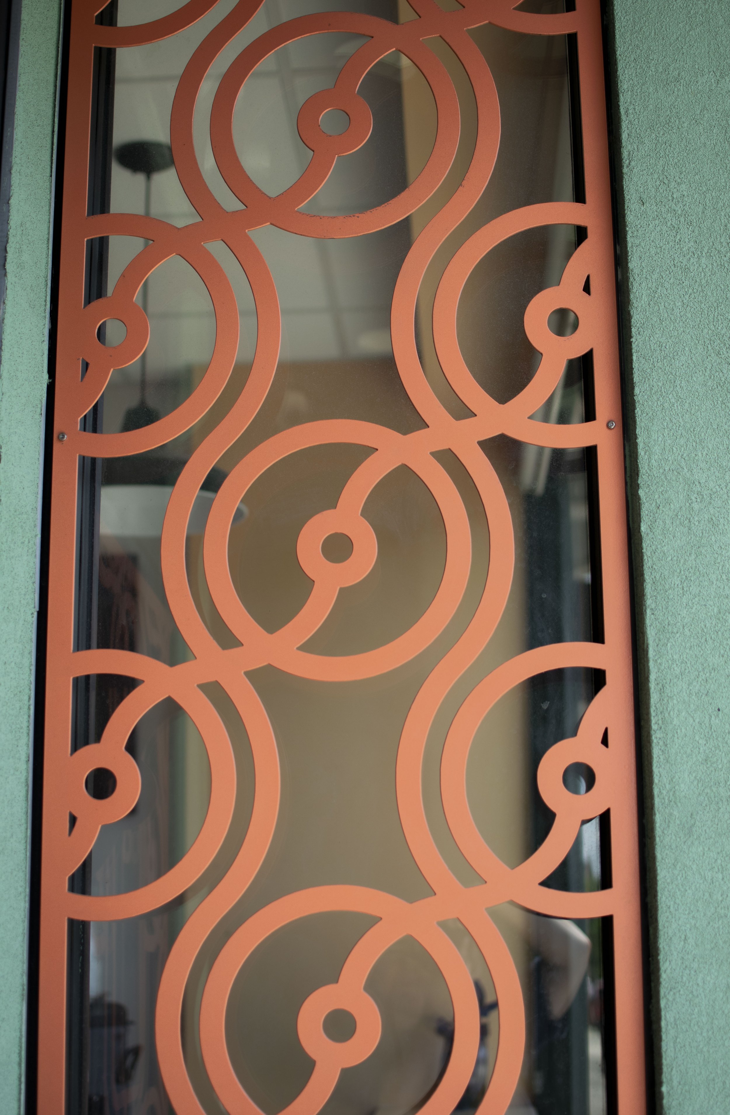 A decorative orange metal screen with interconnected circular and semi-circular patterns, mounted on a wall with green textured paint. Through the screen, a room with a white ceiling, a black lamp, and furniture is faintly visible.