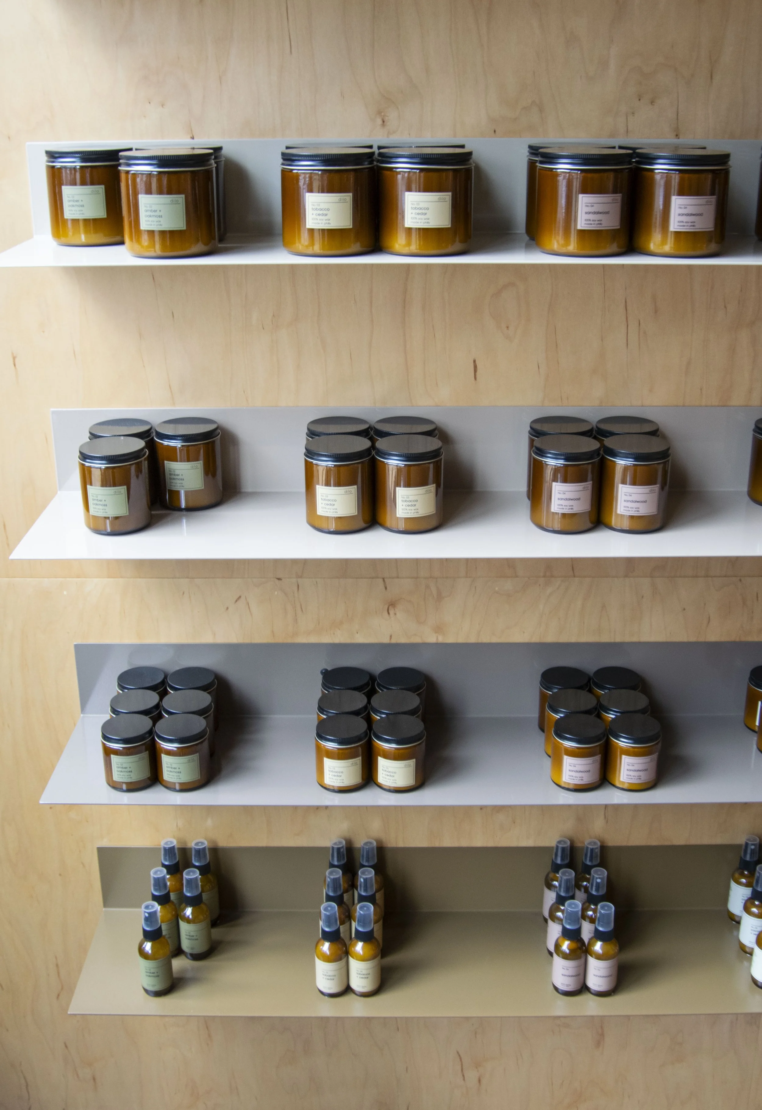 Bent steel shelving holding candles, mounted to plywood walls. Each shelf is a varying shade of beige, creating an overall ombre effect.