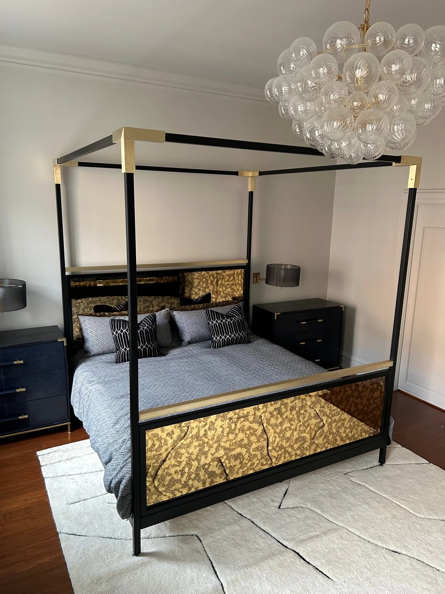 A house bedroom with a bed, black and gold canopy frame, four black and white pillows, two dark gray nightstands, and a unique chandelier with multiple glass globes hanging from the ceiling. The room features off-white walls, a white and black patter