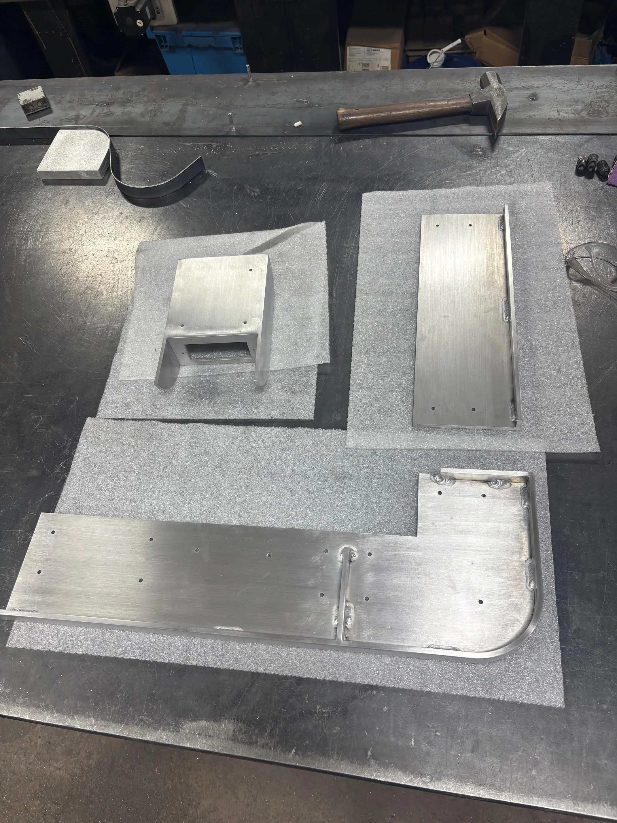 Metalwork workshop with aluminum parts, a hammer, and foam padding on a workbench.