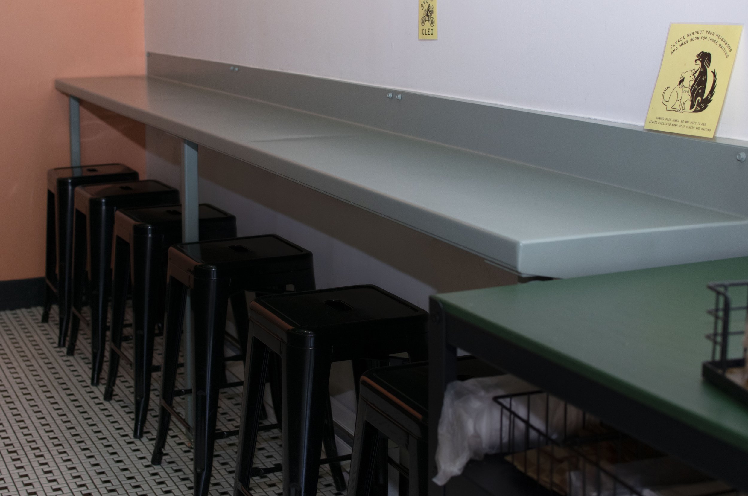 A long white shelf on a wall with yellow signs attached, one with a dog illustration and the other with a dog and cat illustration, next to a green table and black stools, with a black-and-white patterned floor.