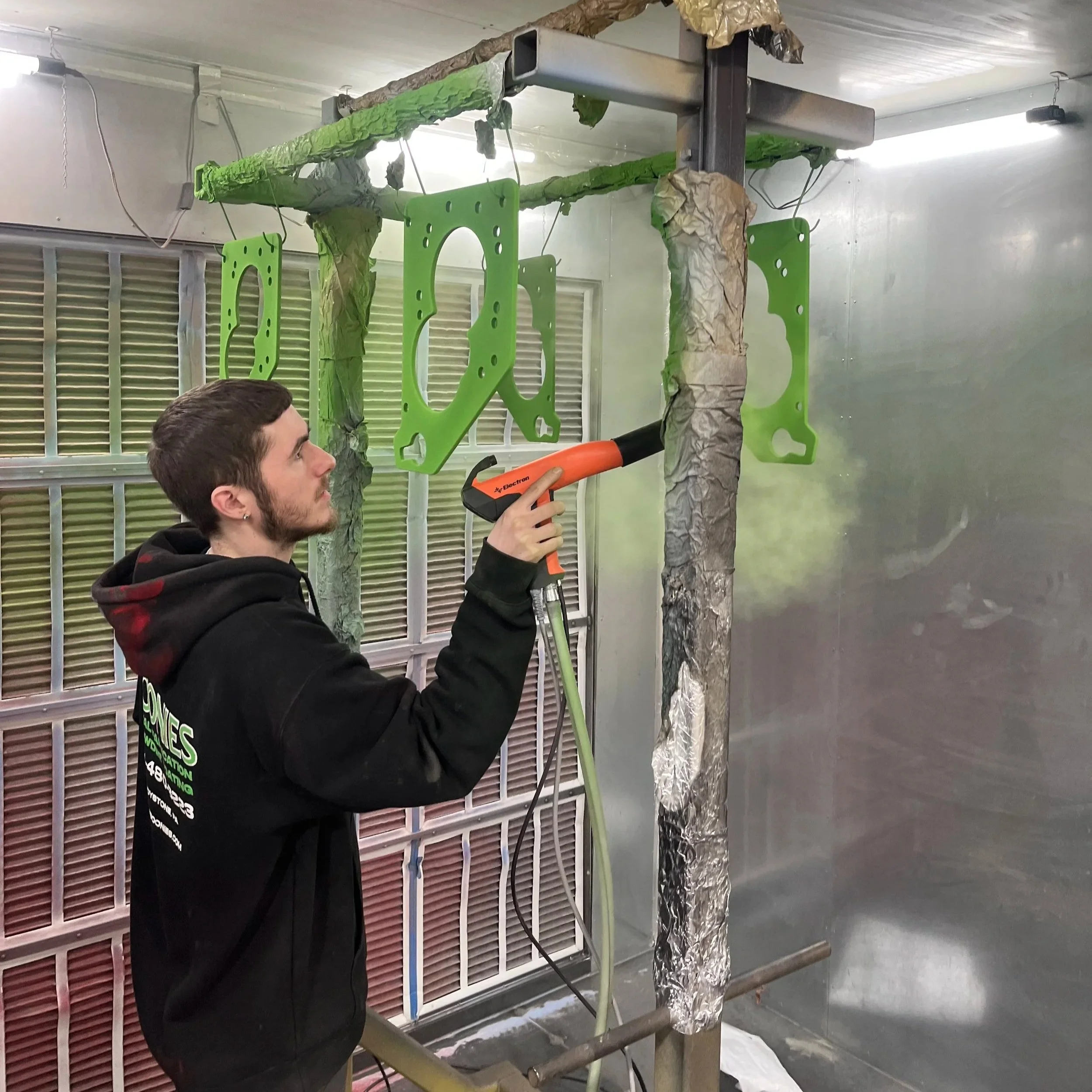 A person is using a heat gun to dry a green automotive part on a makeshift painting station with crumpled paper covering some parts and a metal frame structure in a garage or workshop.