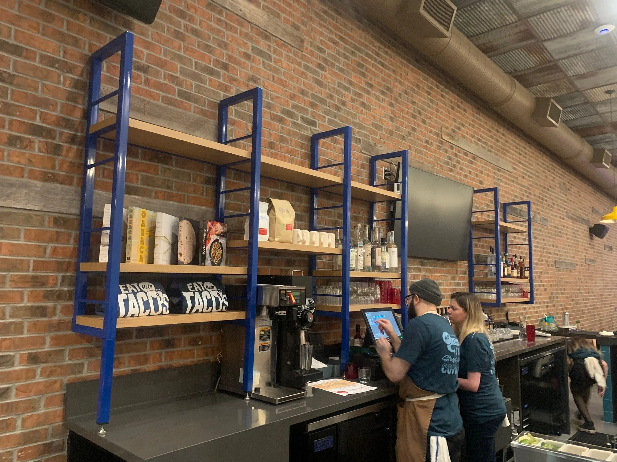 Indoor scene showing a brick wall and steel shelving filled with books, bottles, and boxes. Two people are working at a counter, one using a touchscreen device and the other talking to her. There are signs that read 'Eat Taco' on the counter.