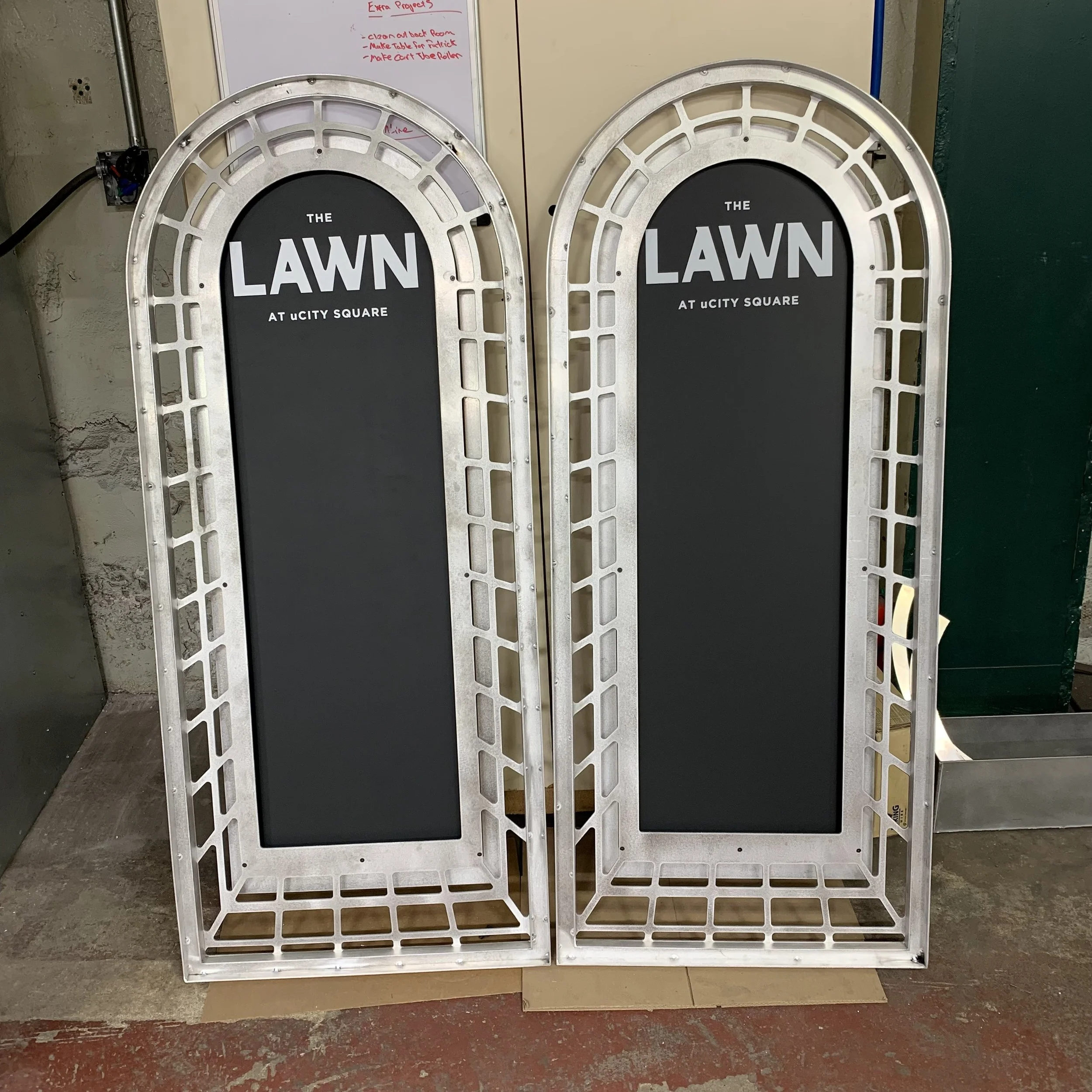 Two large decorative metallic frames with black signs inside reading 'The LAWN at uCIT Y SQUARE'. They are placed on a concrete floor, with a whiteboard and a green wall in the background.