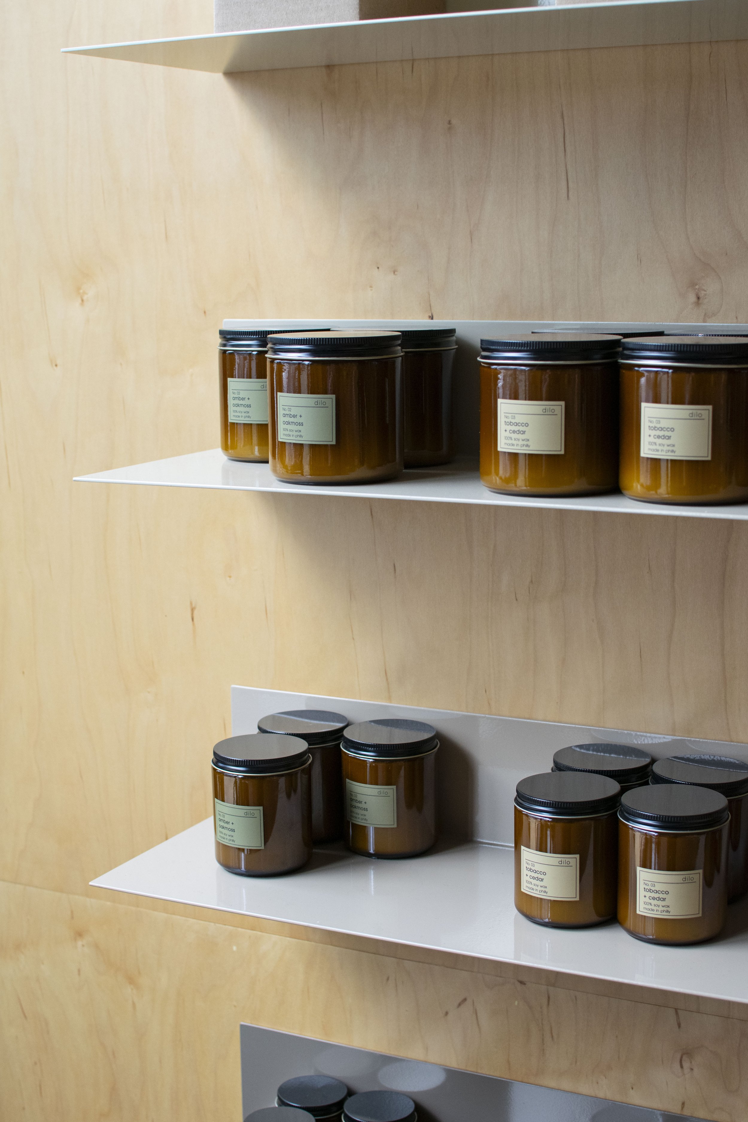 Bent steel shelving holding candles, mounted to plywood walls. Each shelf is a varying shade of beige, creating an overall ombre effect.