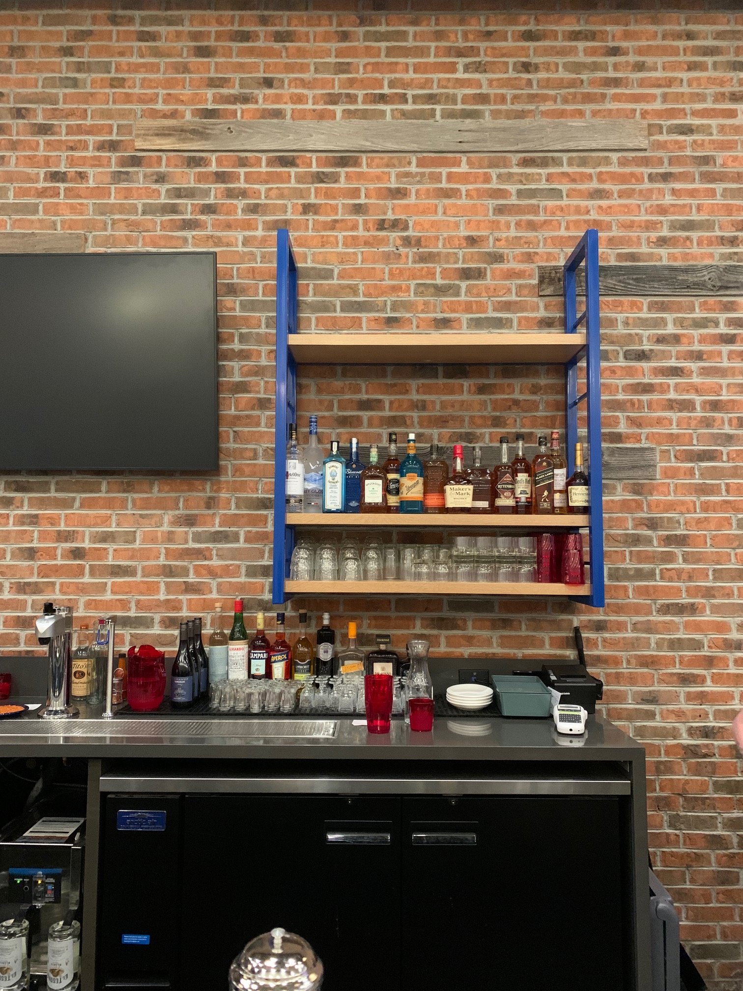 Bar with various bottles of alcohol on a wooden shelf against a brick wall, with glassware and bar tools on a black counter below.