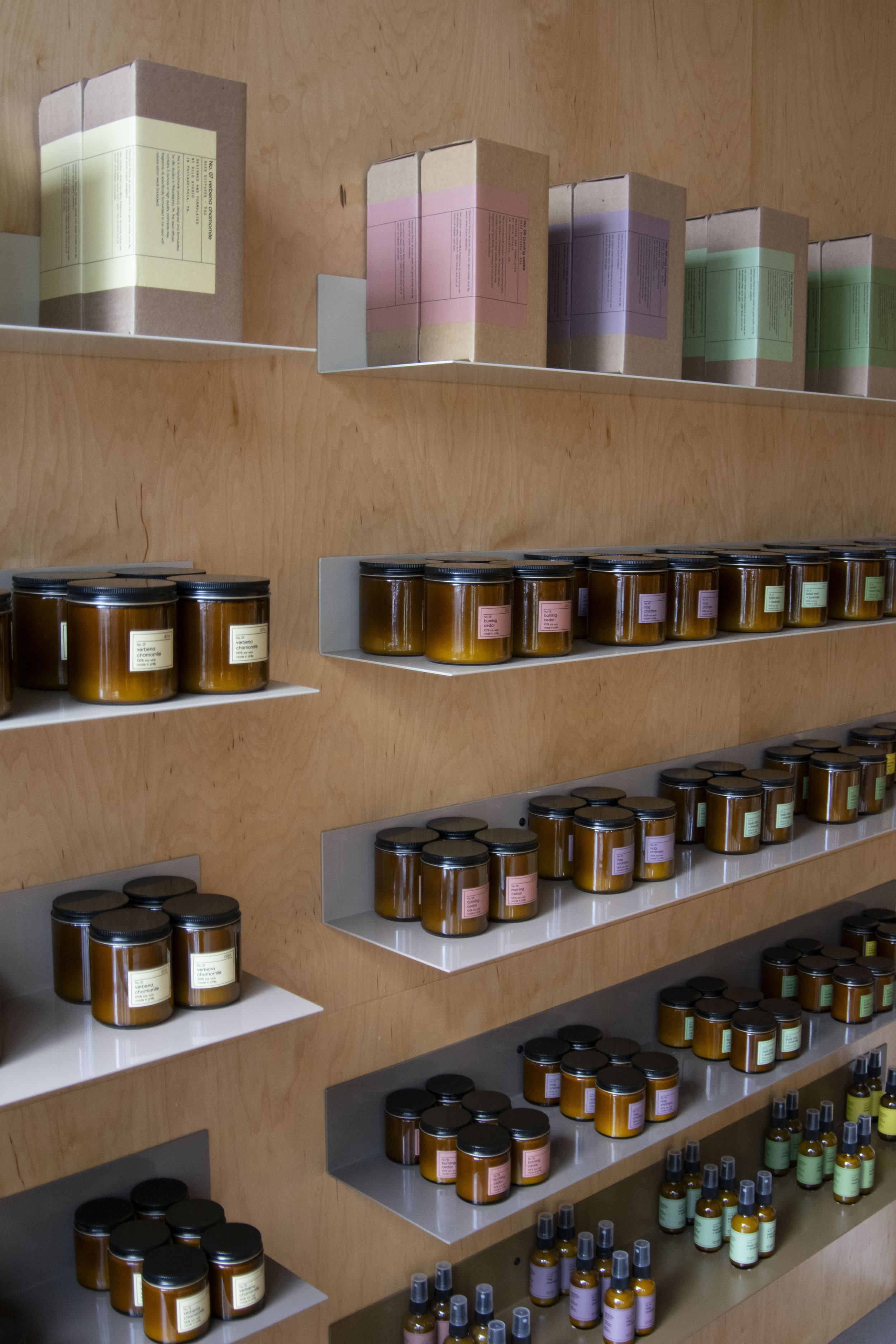 Bent steel shelving holding candles, mounted to plywood walls. Each shelf is a varying shade of beige, creating an overall ombre effect.