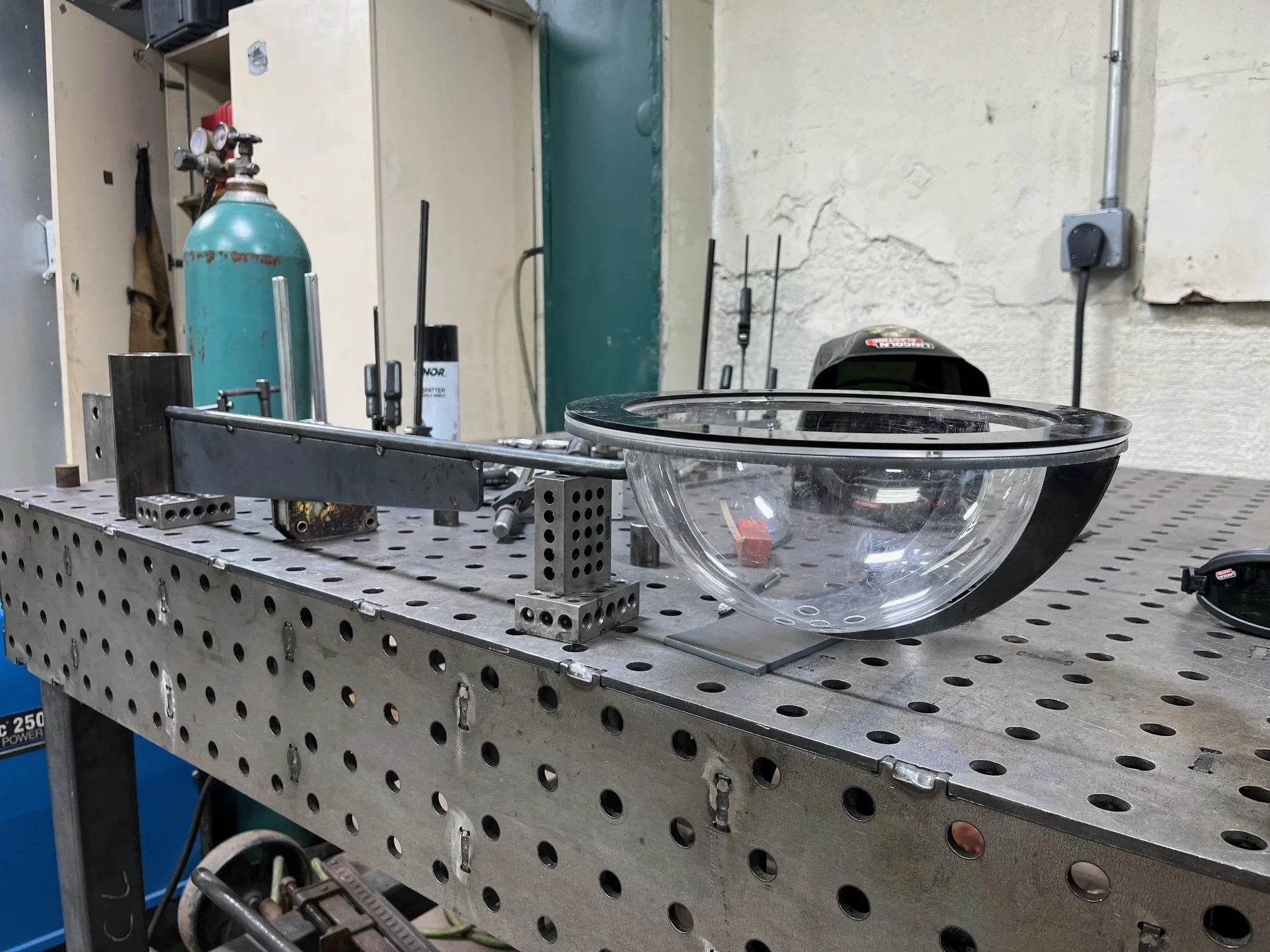 A metal workbench with a glass bowl on top, various tools, metal clamps, and equipment in a workshop with textured beige walls and electrical outlets.