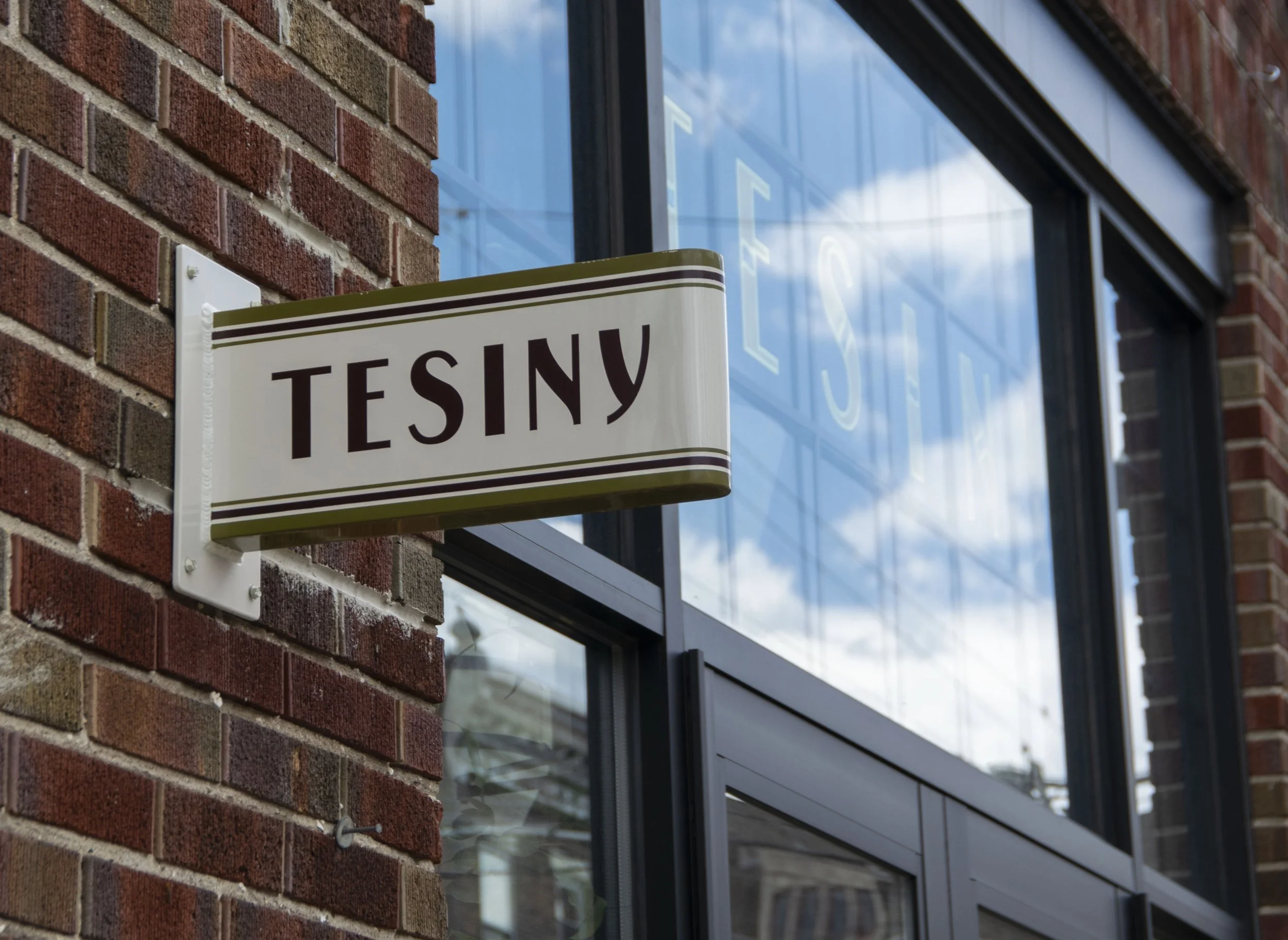 A close-up view of a brick building exterior with a sign that reads 'TESINY' and windows reflecting the sky and clouds.