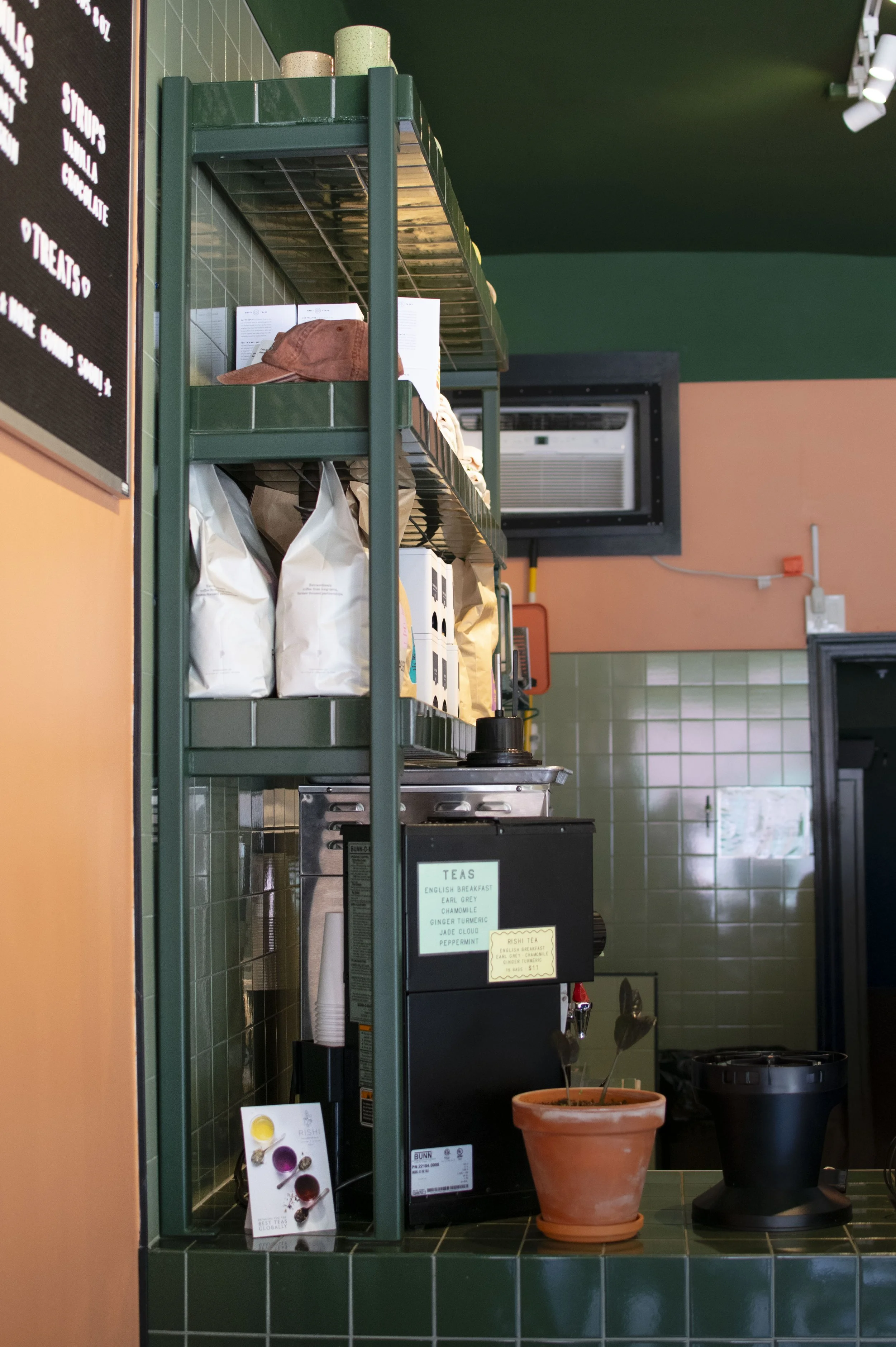Green metal shelf in a café, with various items including a brown baseball cap, bags, and pots on top. Behind the shelf there is a menu board and a window air conditioning unit. In front of the shelf, there is a terracotta pot with a plant and a blac