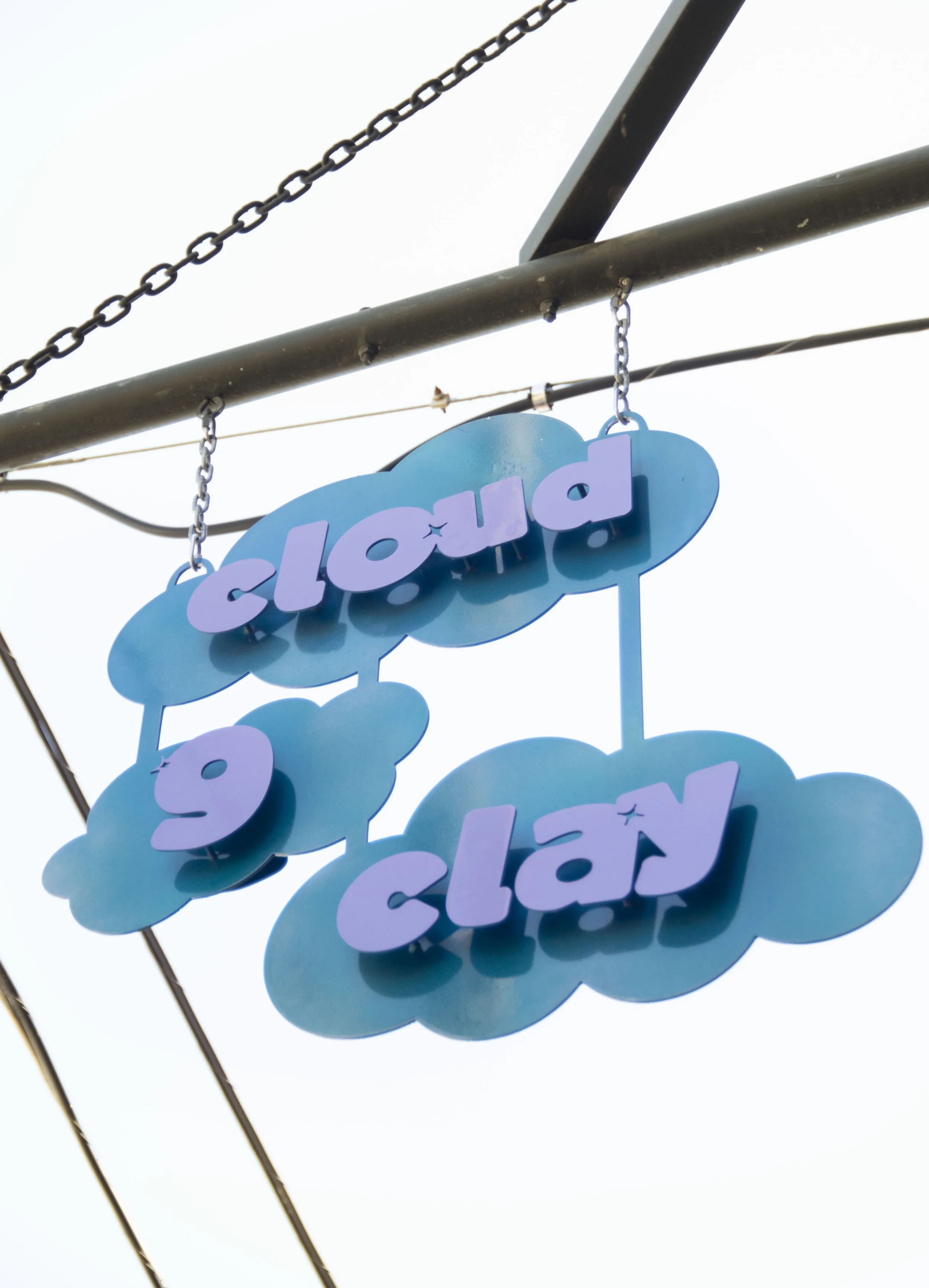Teal cloud-shaped signs hanging from a chain on a metal pole, with the words 'cloud 9 clay' against a bright sky.