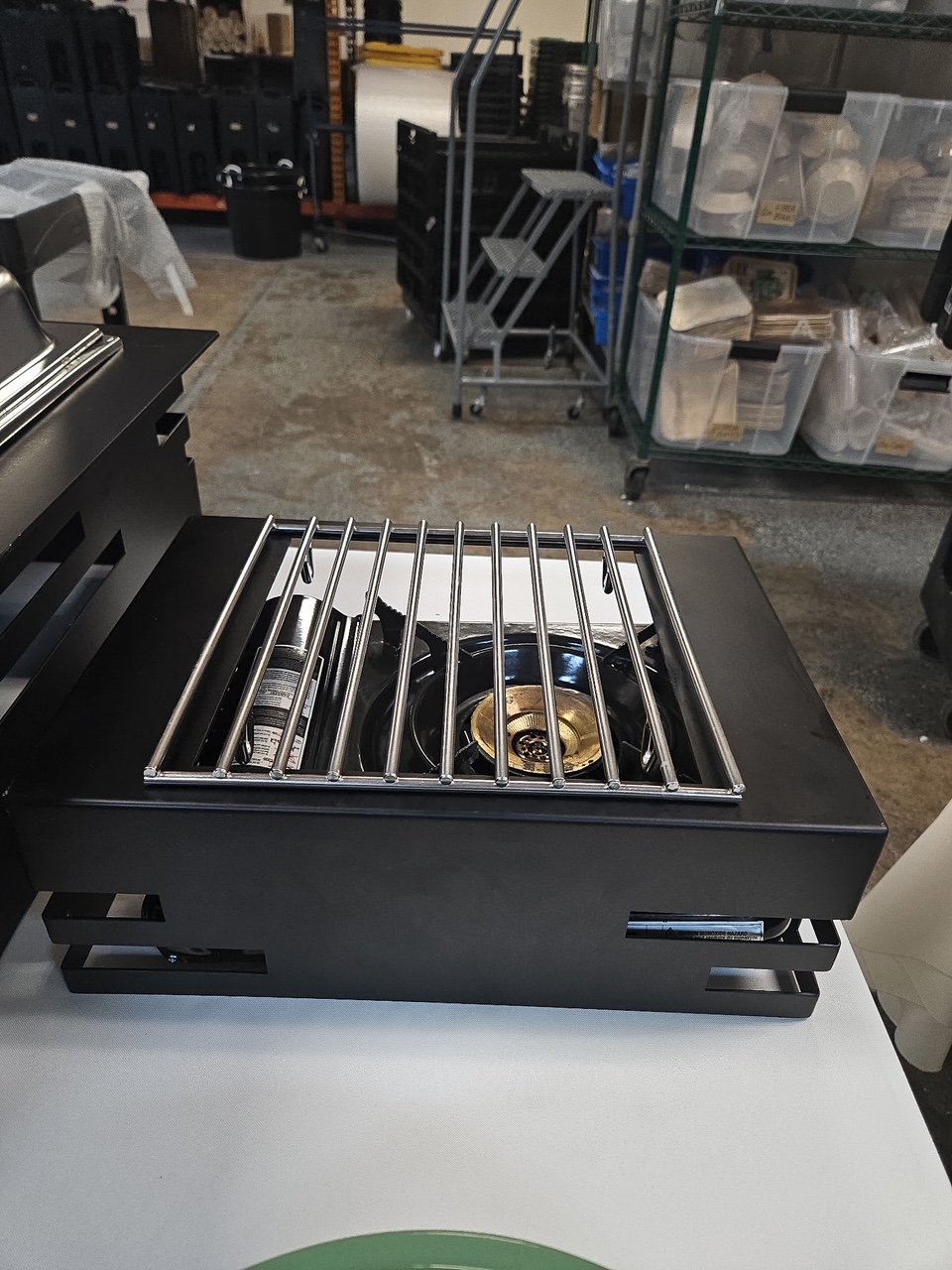 A small black countertop stove with a single gas burner and a metal grate, placed on a white surface in a storage or warehouse area.