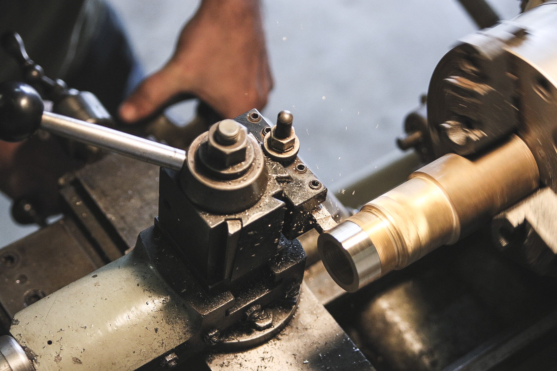 Close-up of a metalworking lathe machine with a worker’s hand adjusting components, as metal shavings and spinning parts indicate metal machining.