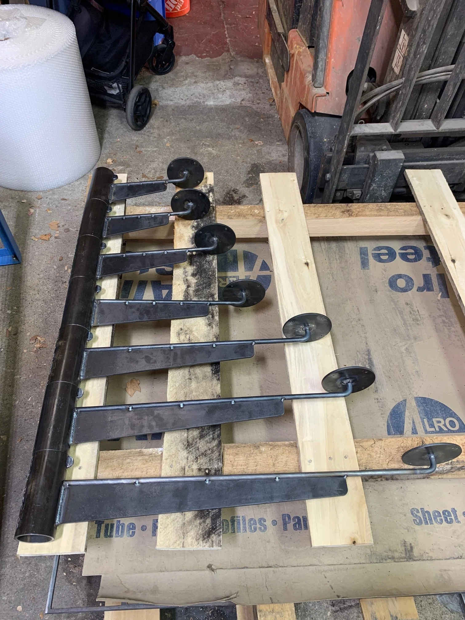 A workshop scene with a partially assembled piece of furniture, possibly a table or workbench, on wooden supports. There are various tools and materials around, including a large white roll of material, a black cart with wheels, and a forklift in the