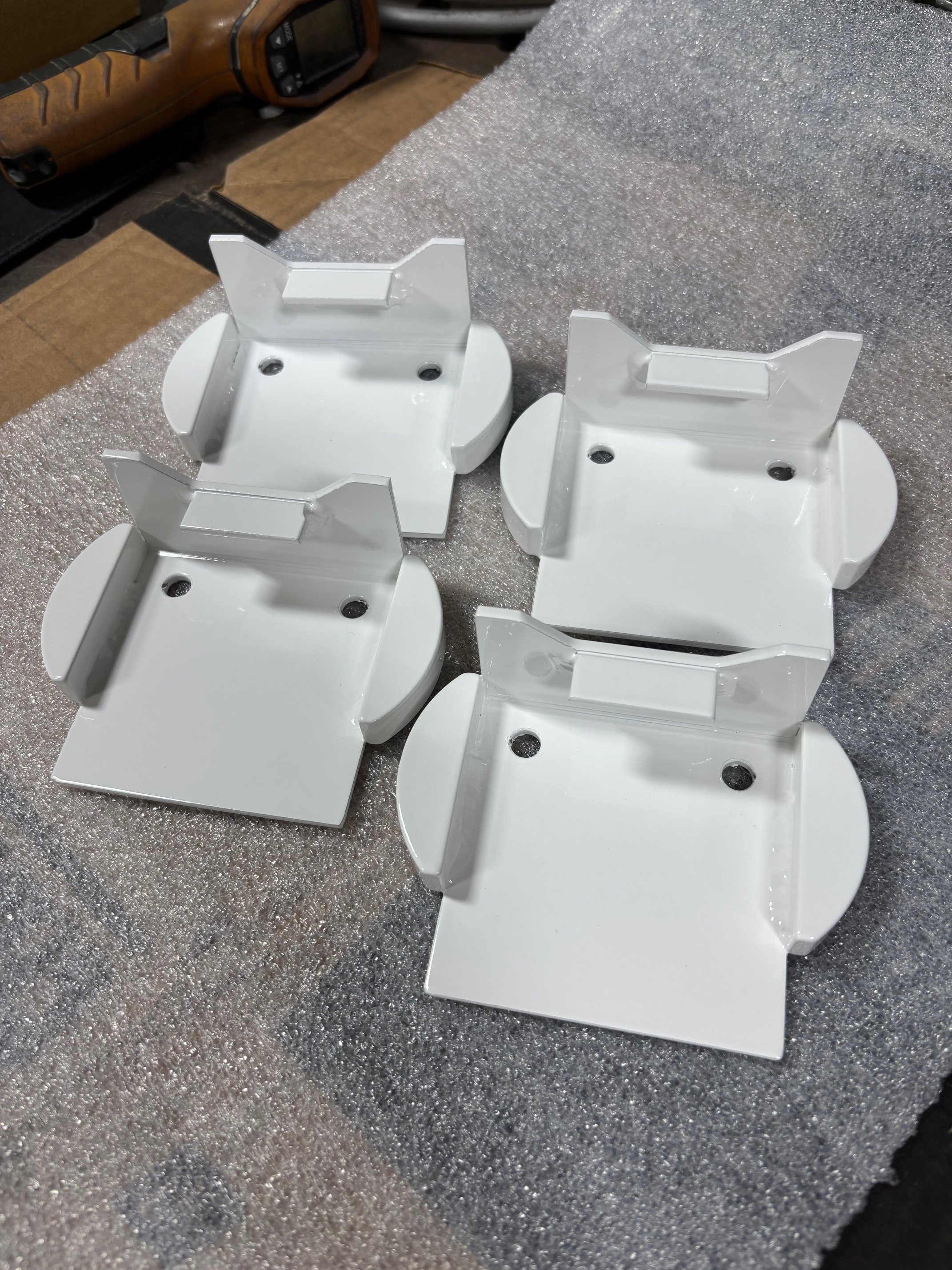 Four white metal brackets with rounded bases and mounting holes on a textured gray surface, with a brown toolbox in the background.