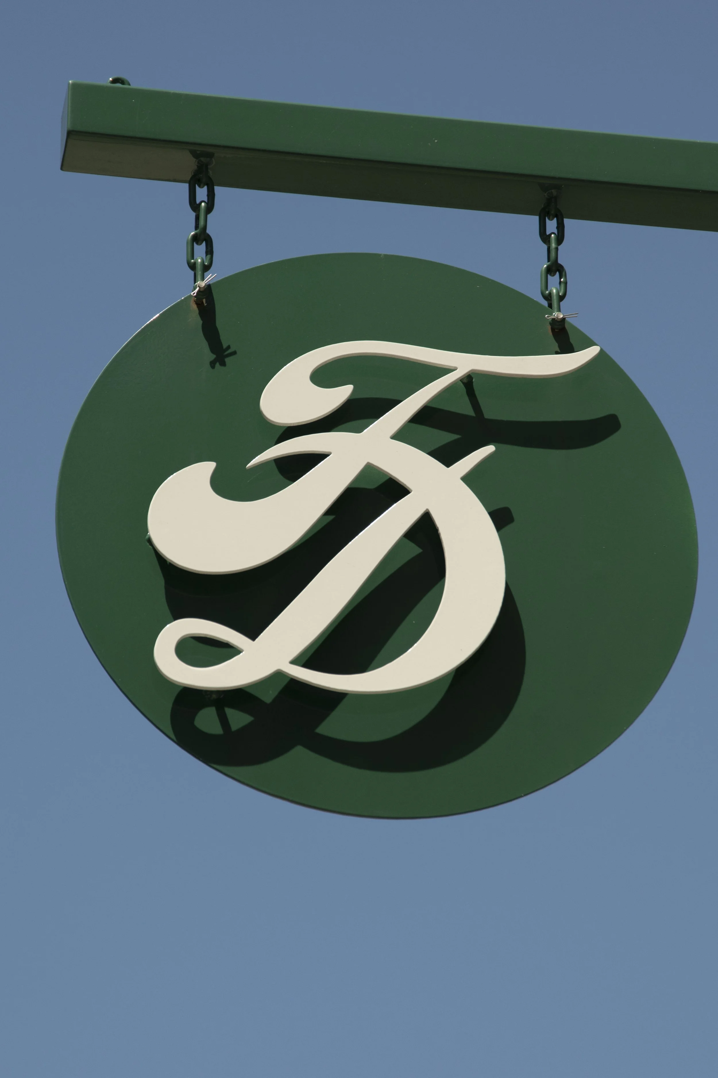 Close-up of a green circular sign with white cursive text, hanging from green metal chains against a clear blue sky.