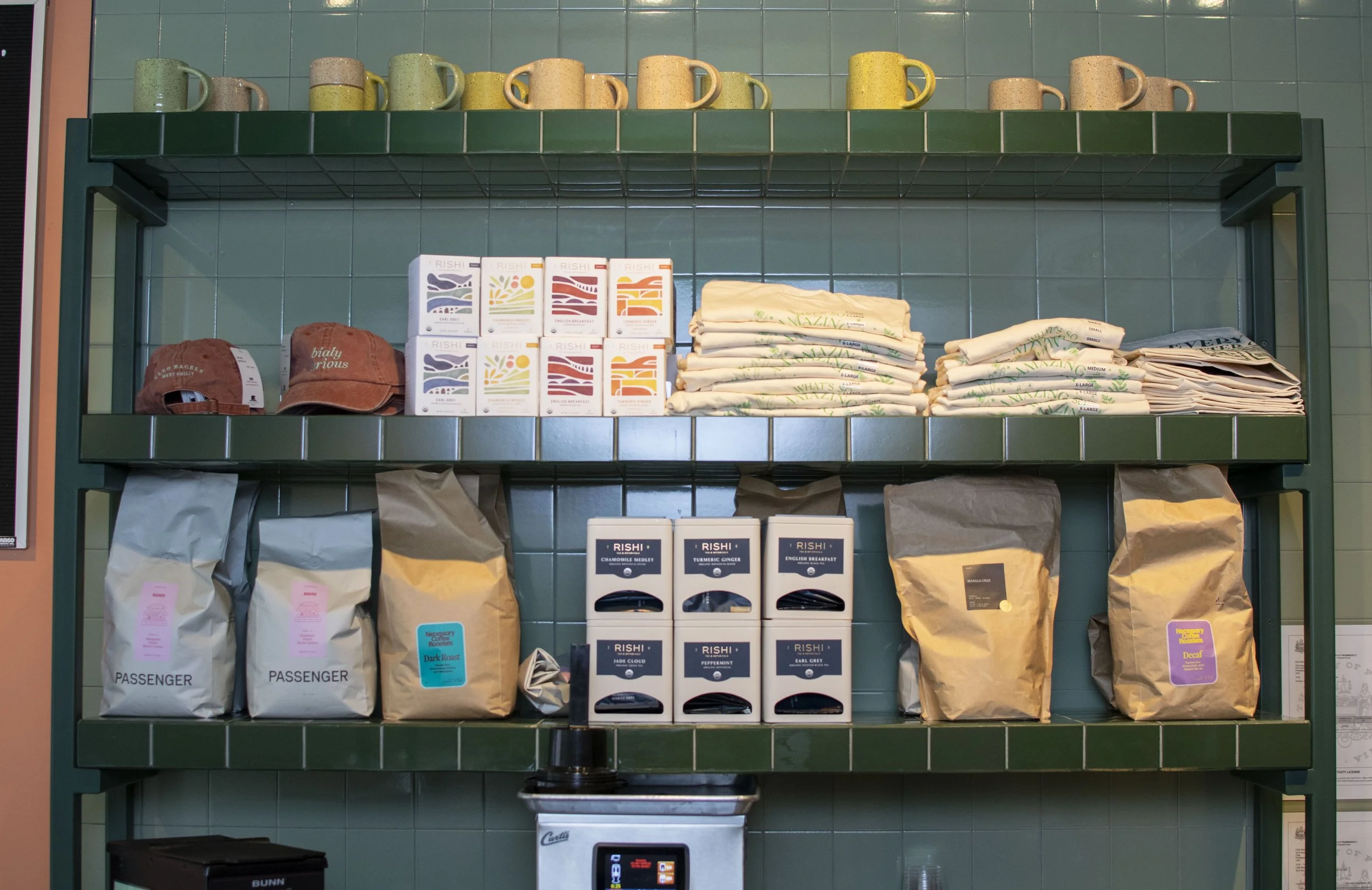 Green metal shelf with coffee mugs on the top row, boxed coffee products, folded cloth napkins, bags of coffee beans, and coffee accessories on the shelves below.