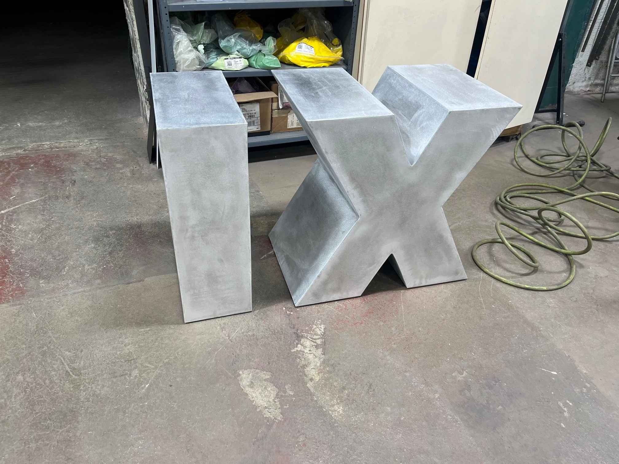 A large silver metallic letter 'V' sculpture placed on a concrete floor. In the background, there is a gray shelving unit with plastic bags, cardboard boxes, and other small items. To the right, some yellow-green electrical cords are coiled on the fl