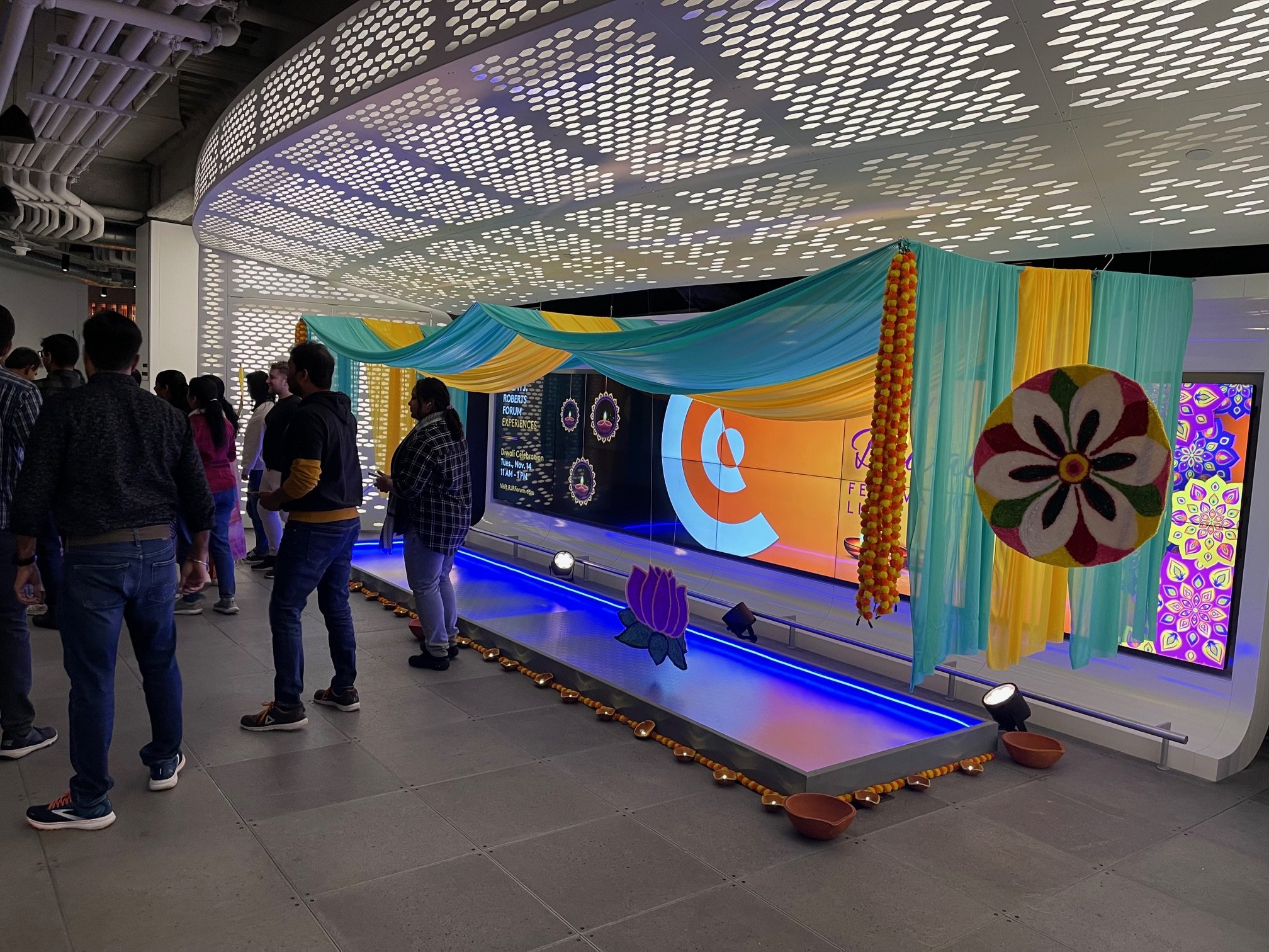 People gathered around a traditional Indian decorative display with colorful drapes, hanging marigold flower garlands, floral decorations, and a large circular floral ornament at an indoor event.