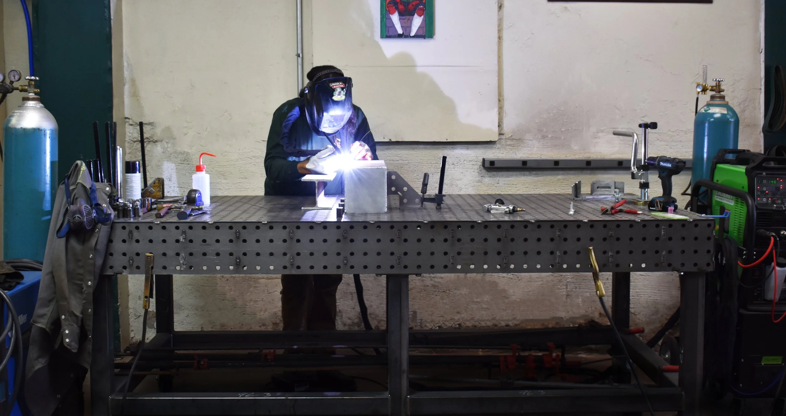 A person wearing a welding helmet working on a metal project at a workshop table with tools and welding equipment around.