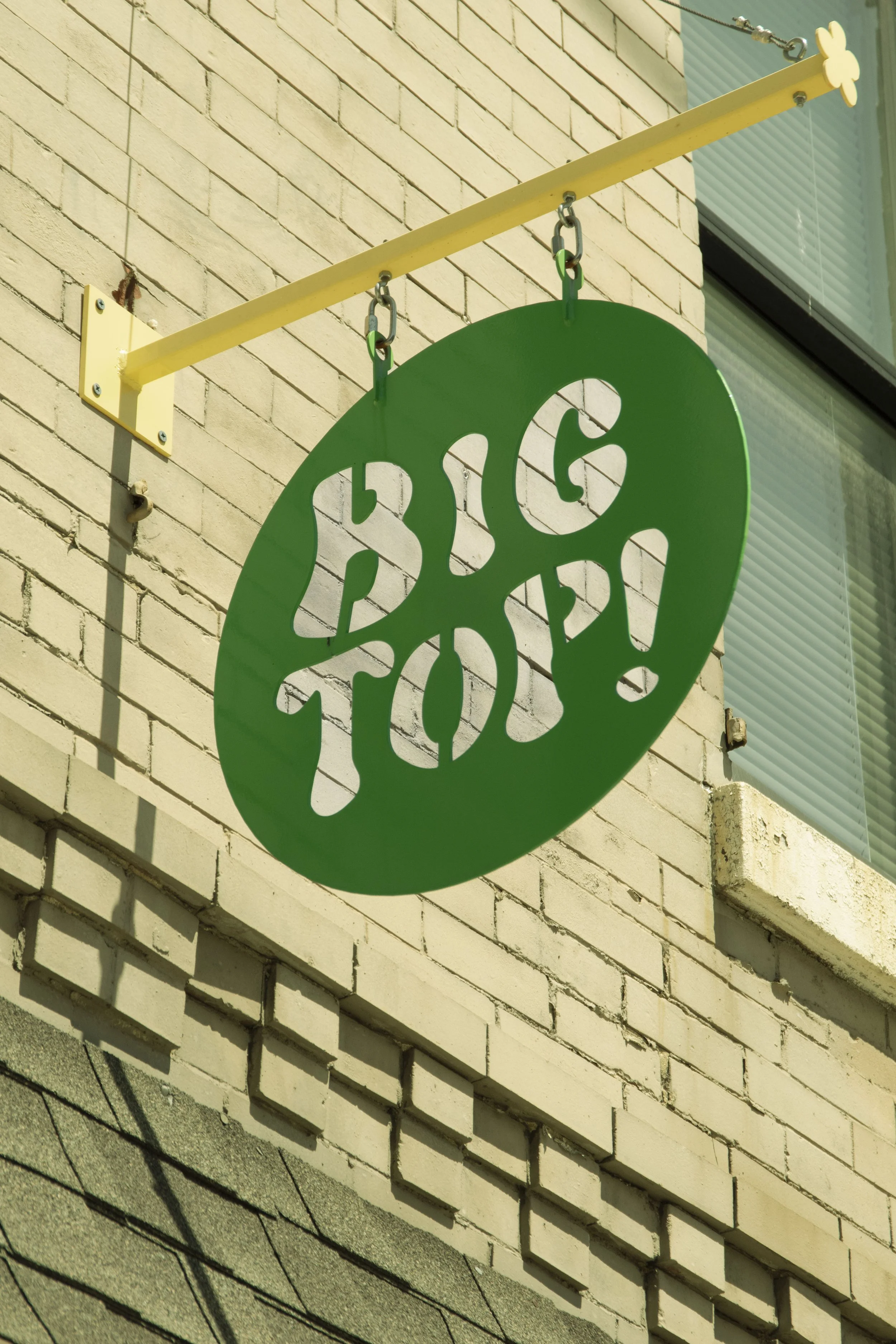 A green decorative sign hanging from a yellow bracket on an exterior brick wall of a building, with part of a window visible in the background.