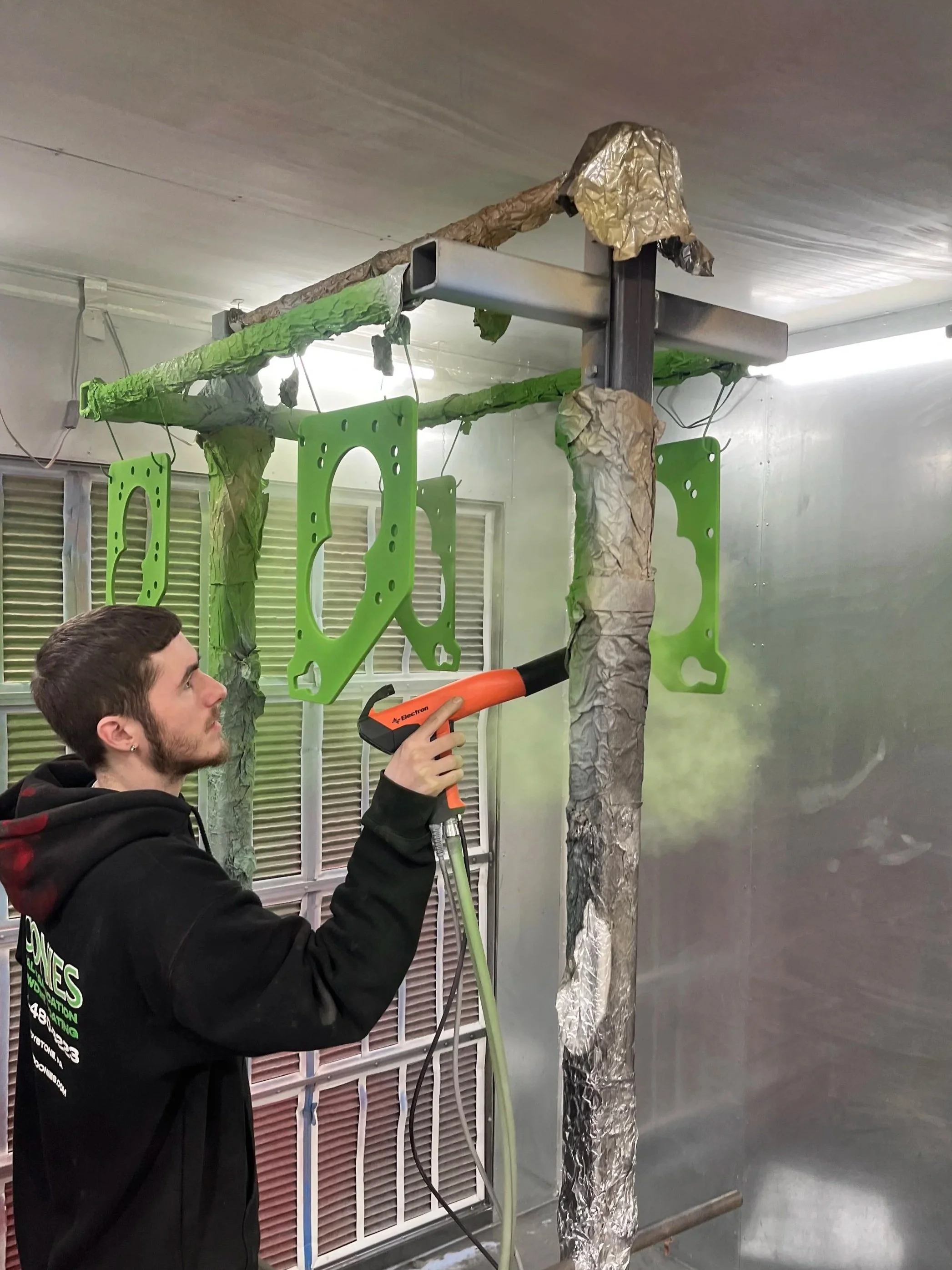 A person using a heat gun on a green gasket attached to a metal frame that is wrapped in foil and paper, in an industrial or workshop setting.