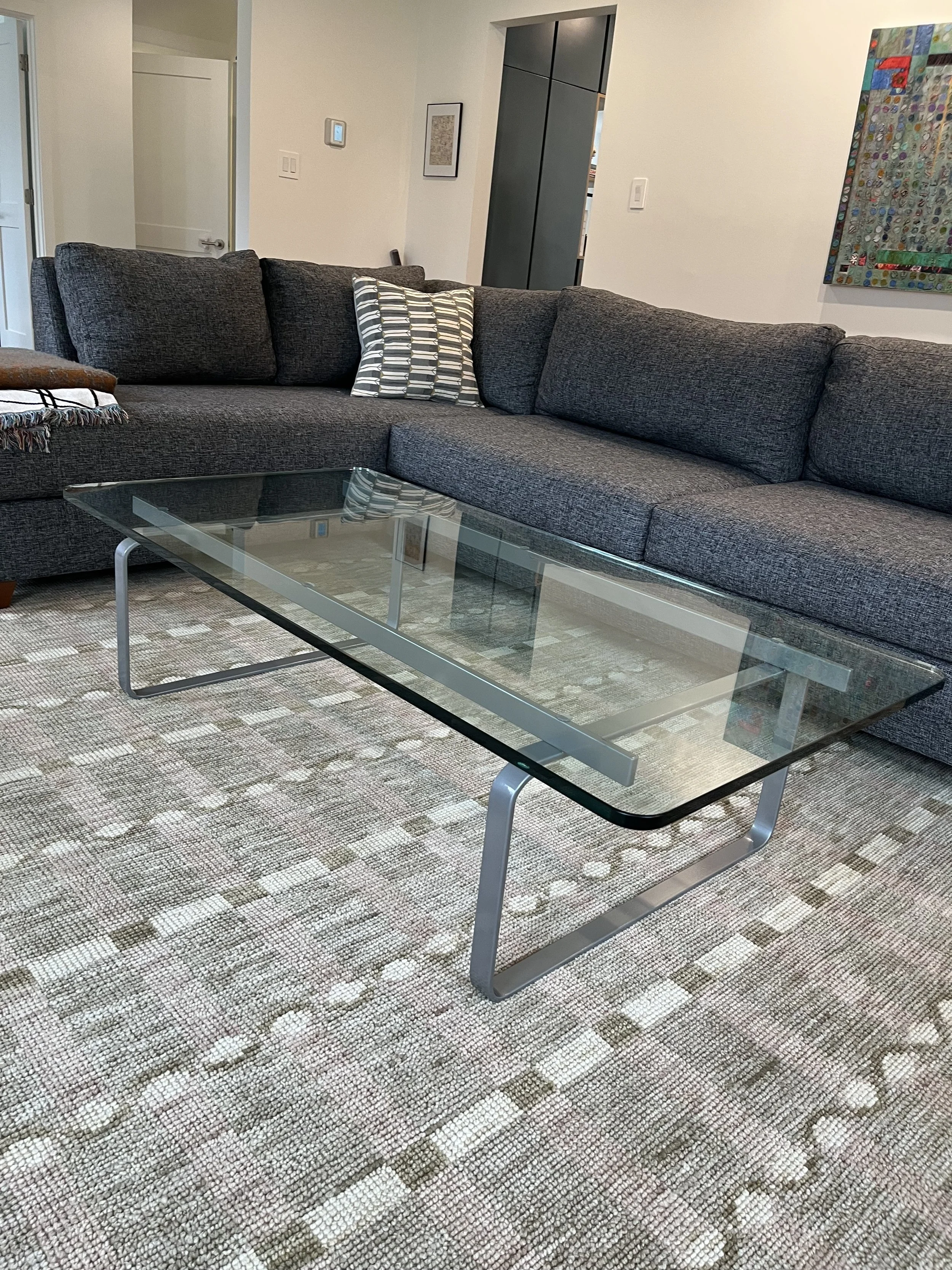 A living room with a dark gray sectional sofa, a glass coffee table, a patterned rug, and wall art. There is a throw blanket on the arm of the sofa and an accent pillow on the sofa.