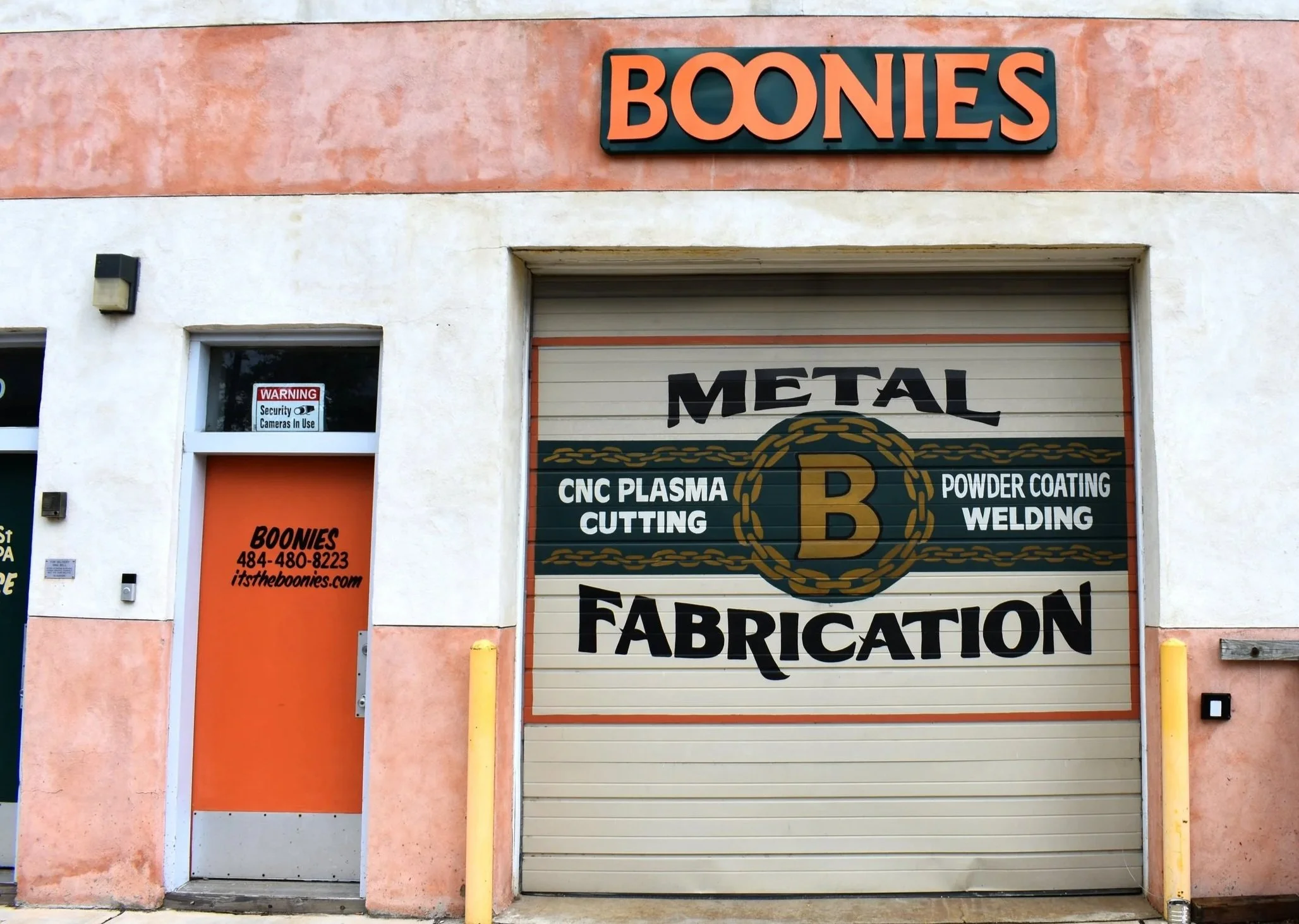 Exterior of a building with a sign that reads 'BOONIES' above a door, and a large closed metal door with text promoting metal fabrication services, including CNC plasma cutting, powder coating, welding, and fabrication.