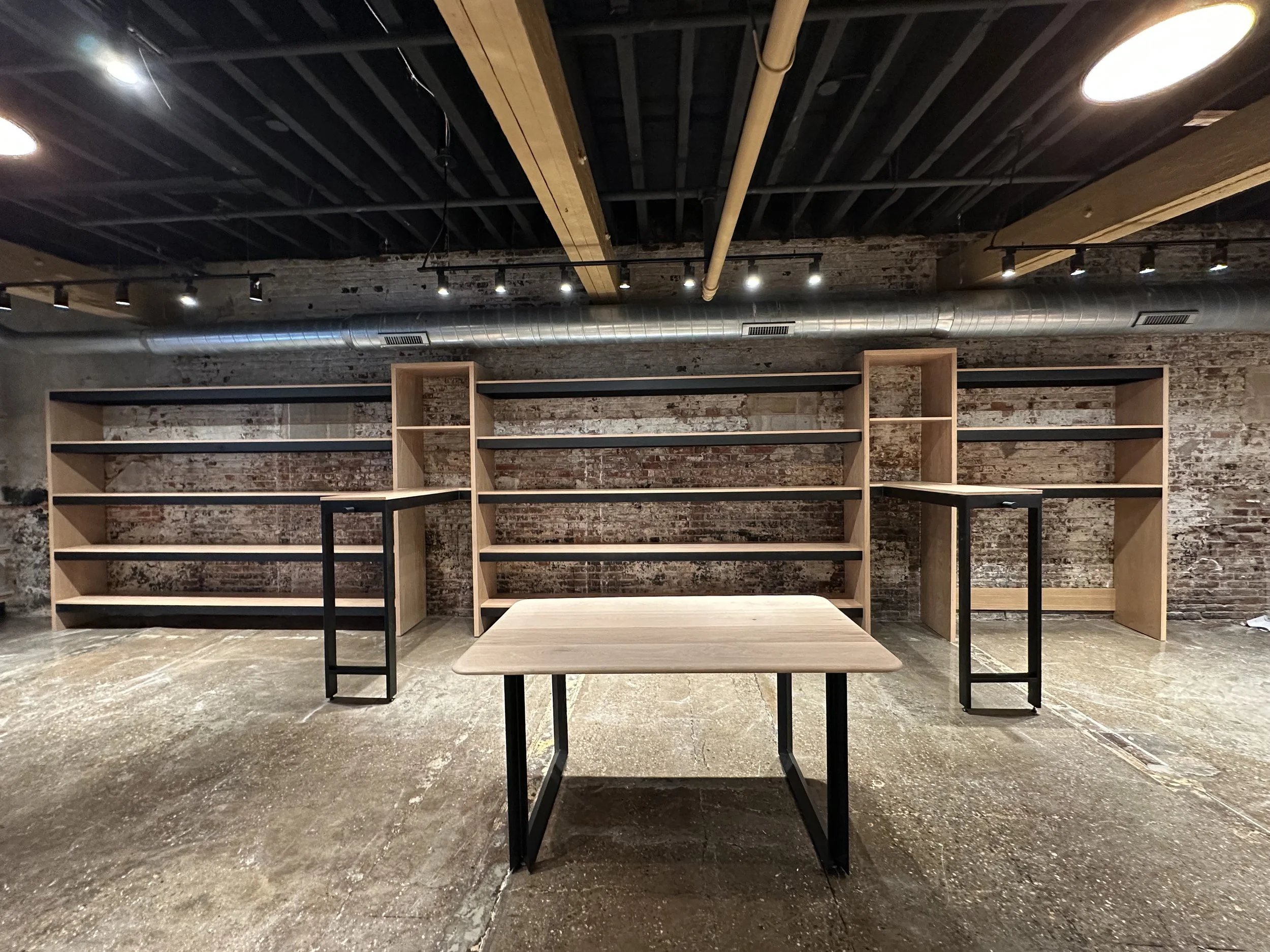 Interior of a modern space with exposed brick walls, black ceiling with wooden beams, track lighting, and metal ductwork. There are wooden bookshelves and tables with black metal legs.
