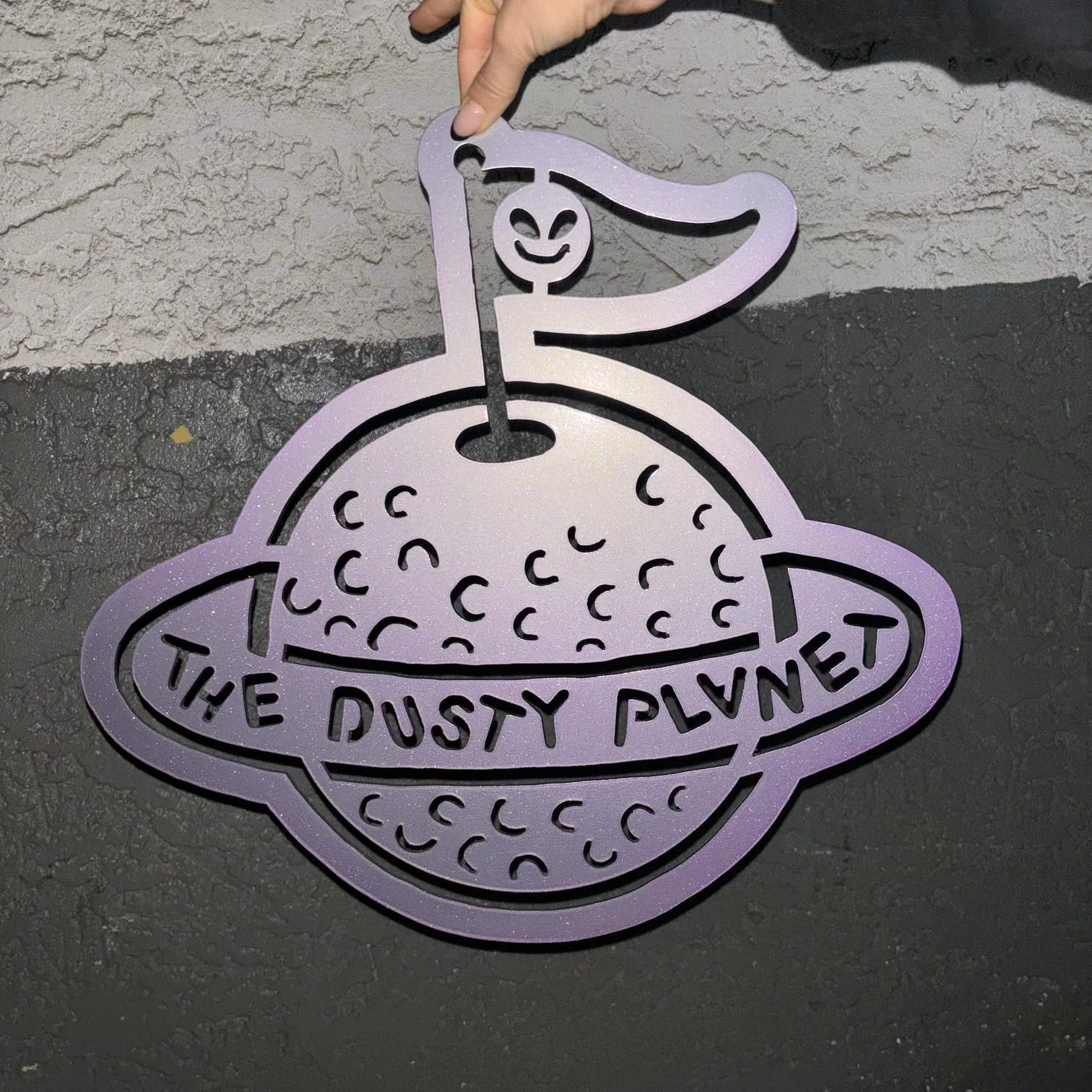 A purple metal ornament shaped like a slice of pizza with a happy face on a pepper stuck into the pizza. The ornament has the words "The Dusty Planet" written on it.