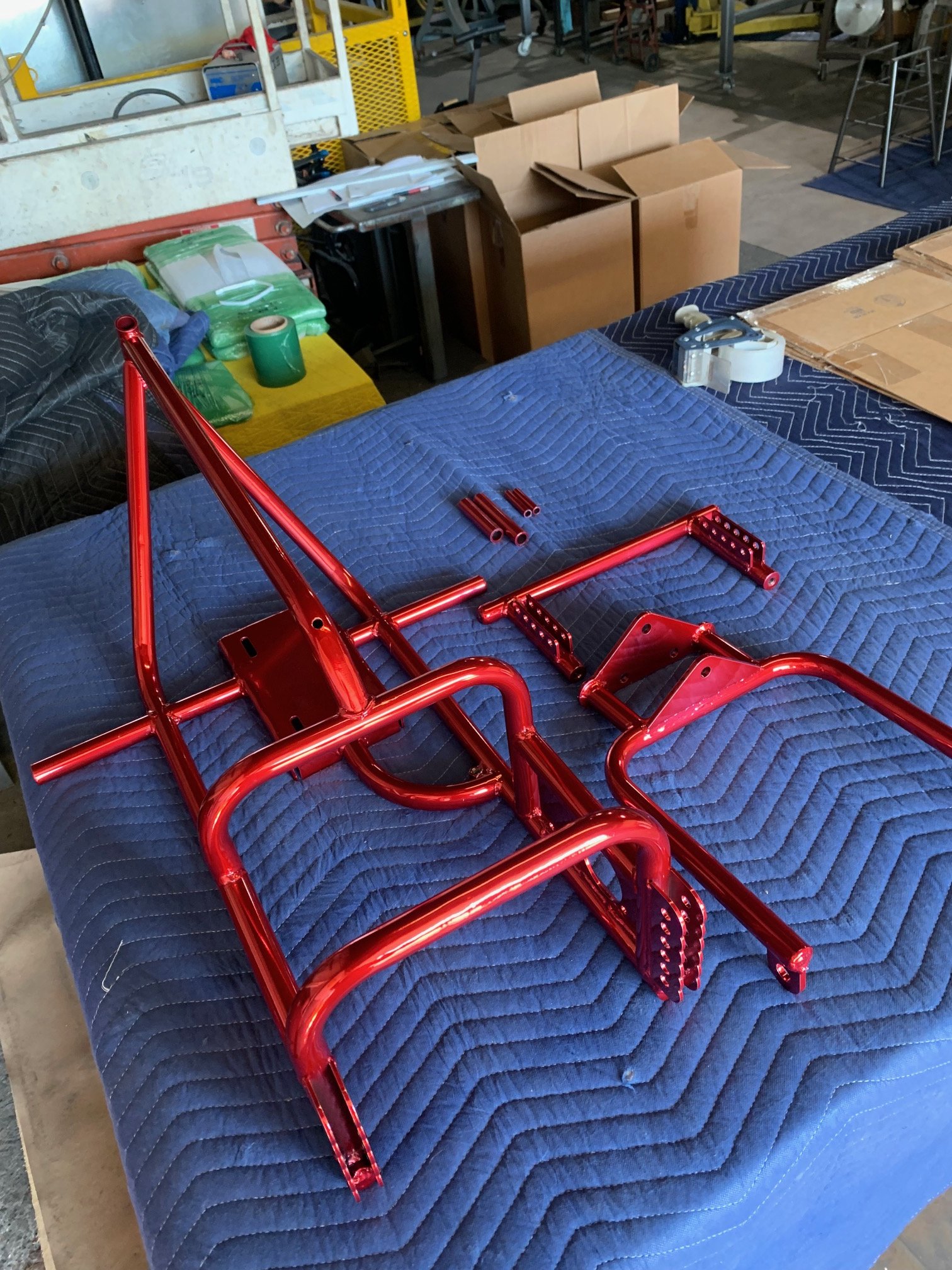 Unassembled red metallic frame parts laid out on a quilted blue blanket in a workshop setting with cardboard boxes and tools in the background.