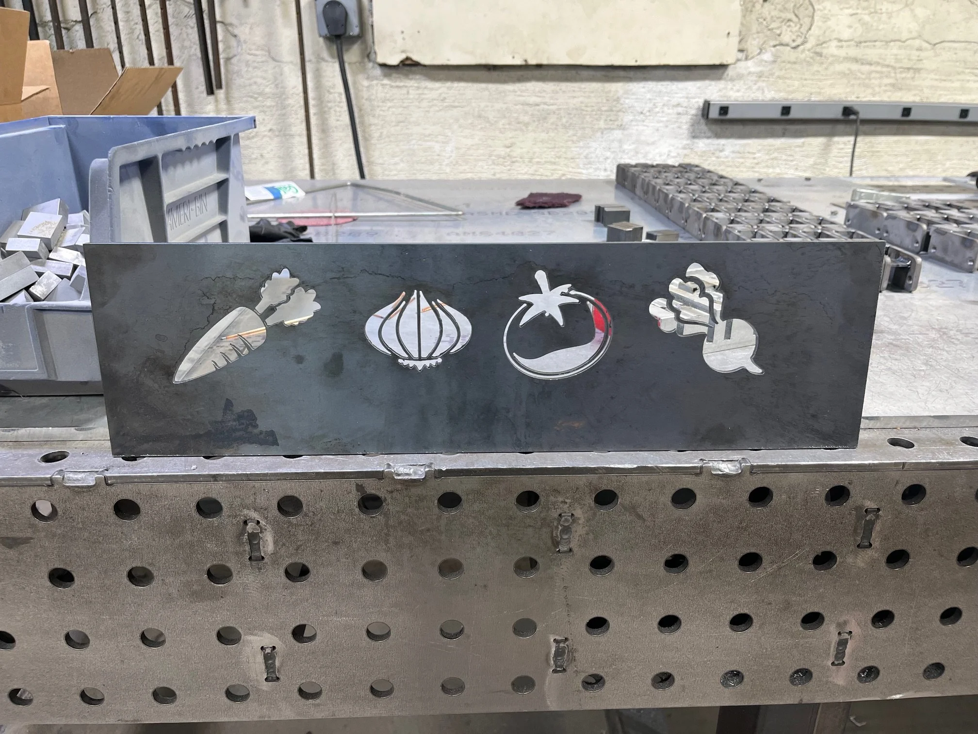 A metal workbench with various tools and small metal parts, including wire mesh trays and cutouts of vegetables like a carrot, onion, tomato, and bell pepper, placed on and around the workbench.
