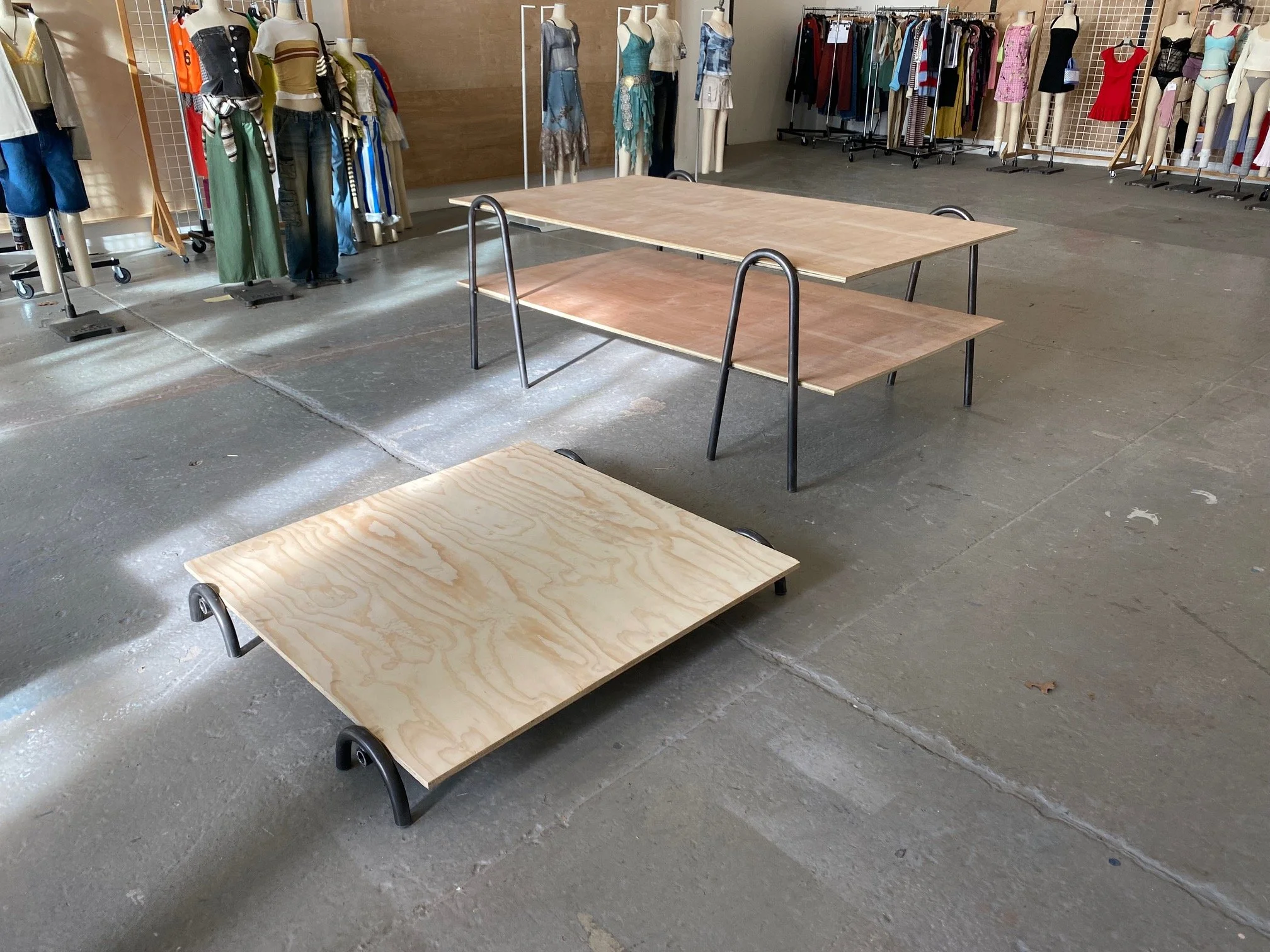 Empty retail store with clothing mannequins displaying various outfits and clothing racks in the background, and two large wooden table platforms in the foreground.