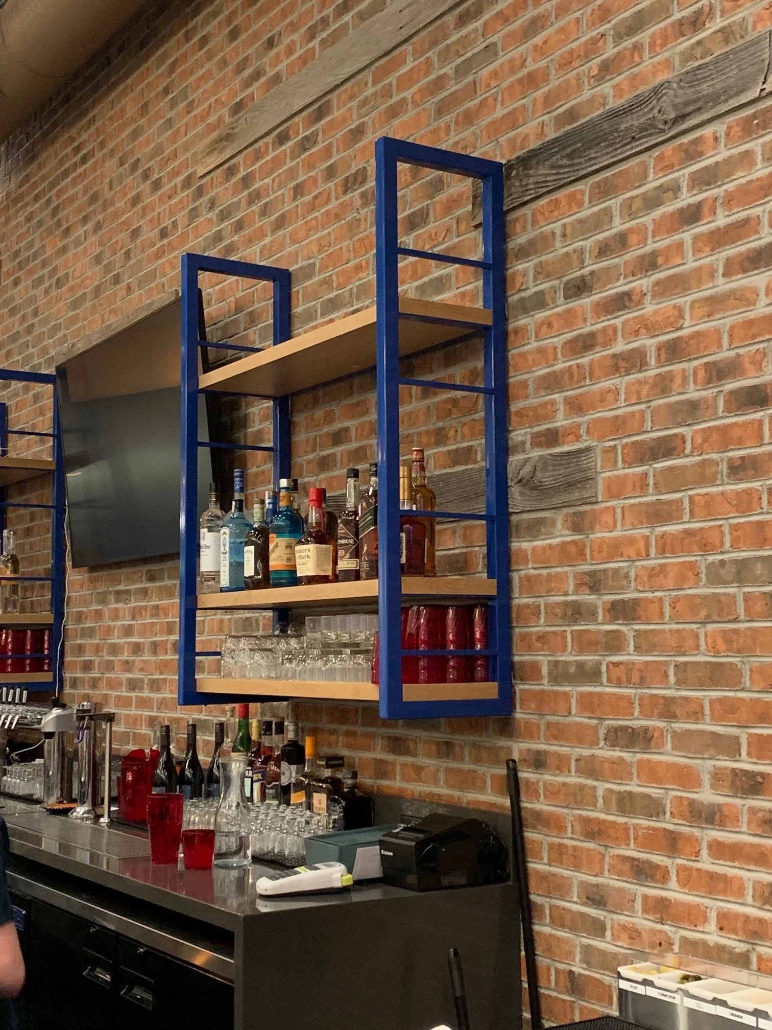 A brick wall with a blue metal and wood shelving unit containing liquor bottles, glasses, and bar accessories. A bar counter in front with various bar tools, a cash register, and containers of garnishes.