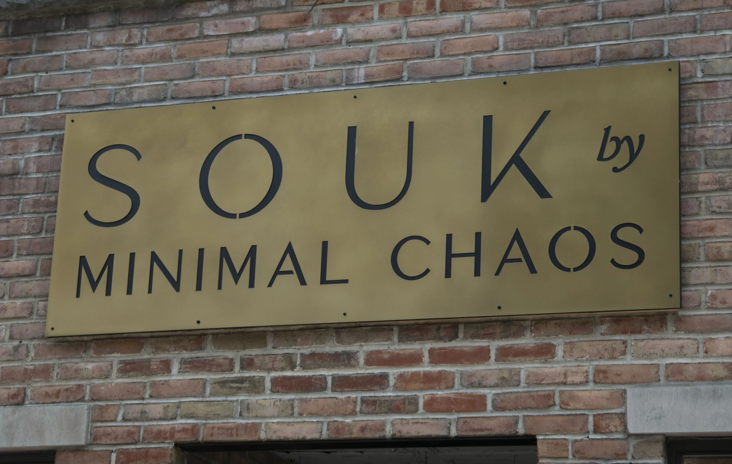 A gold-colored sign with black text reading "SOUK MINIMAL CHAOS" mounted on a red brick wall.