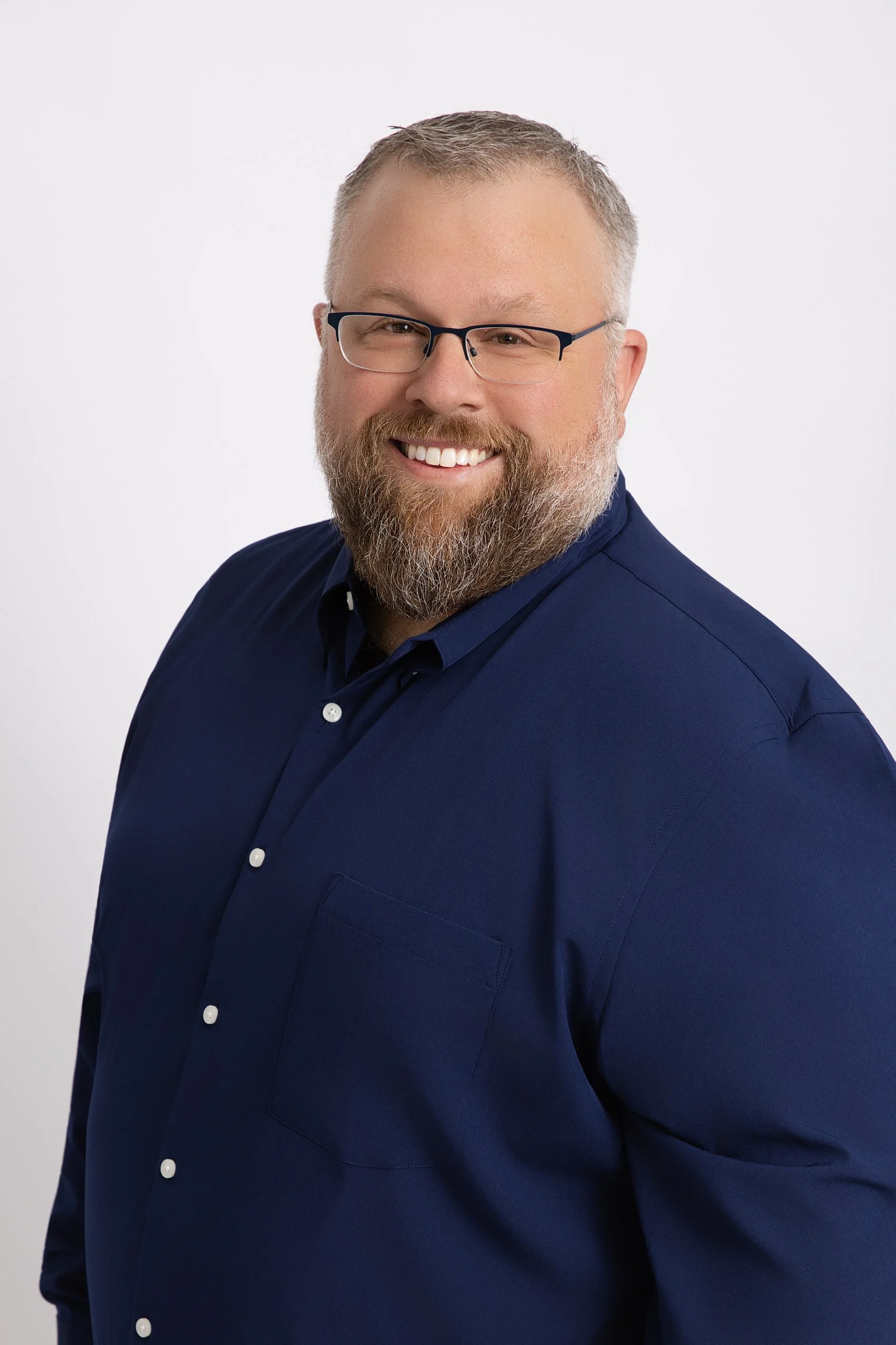 Close-up of a smiling man with glasses, a beard, wearing a dark blue button-up shirt, against a plain white background.