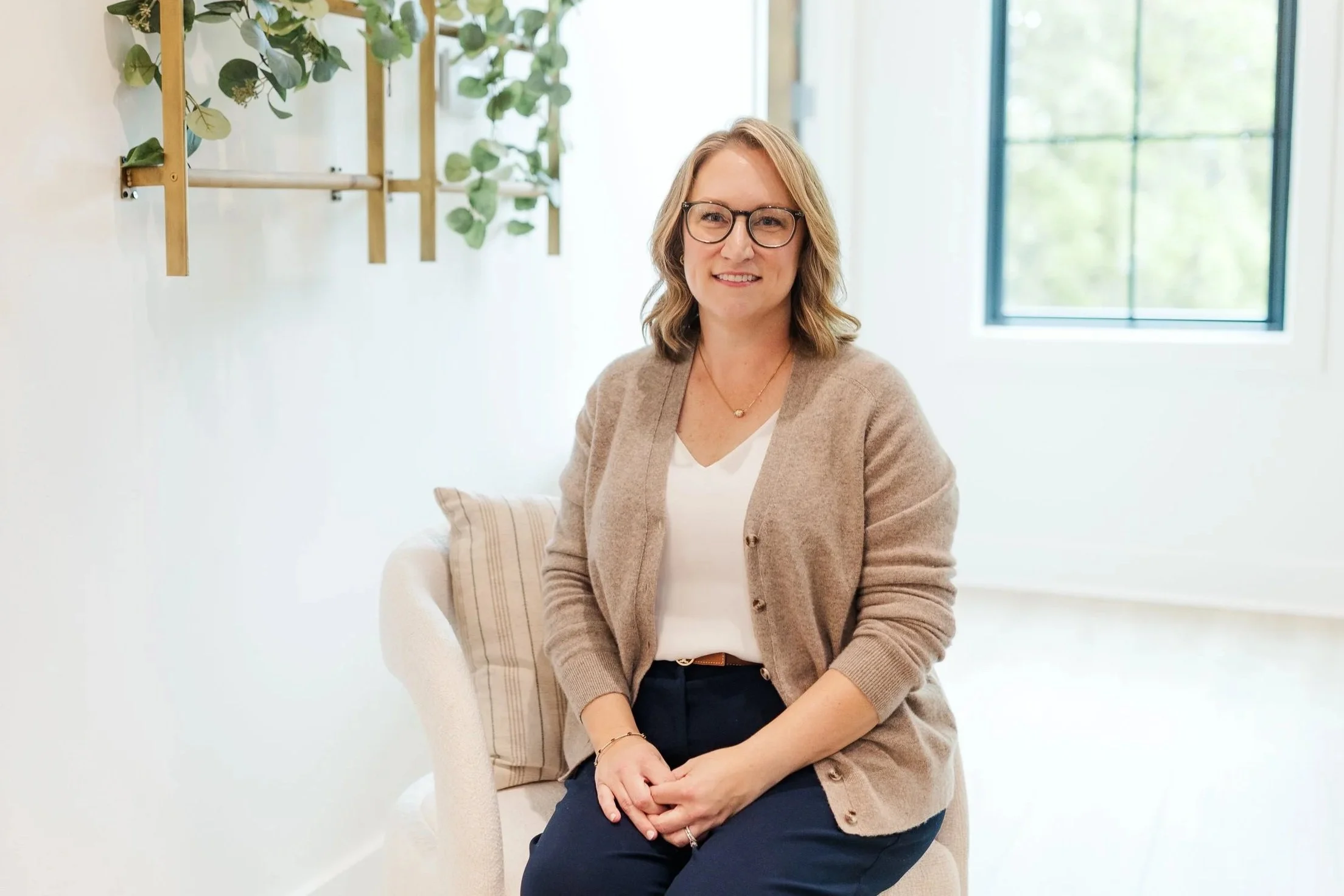 Woman seated in a bright, modern office, smiling at the camera while wearing glasses and business-casual attire, creating a warm and approachable leadership coaching presence.