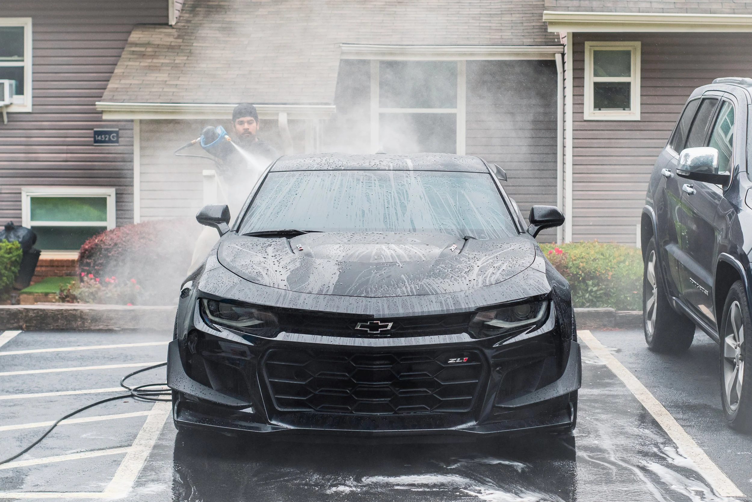 Person pressure washing a black Chevrolet Camaro ZL1 sports car in a parking lot with a hose, with soap and water on the car's surface, and water spray in the air.
