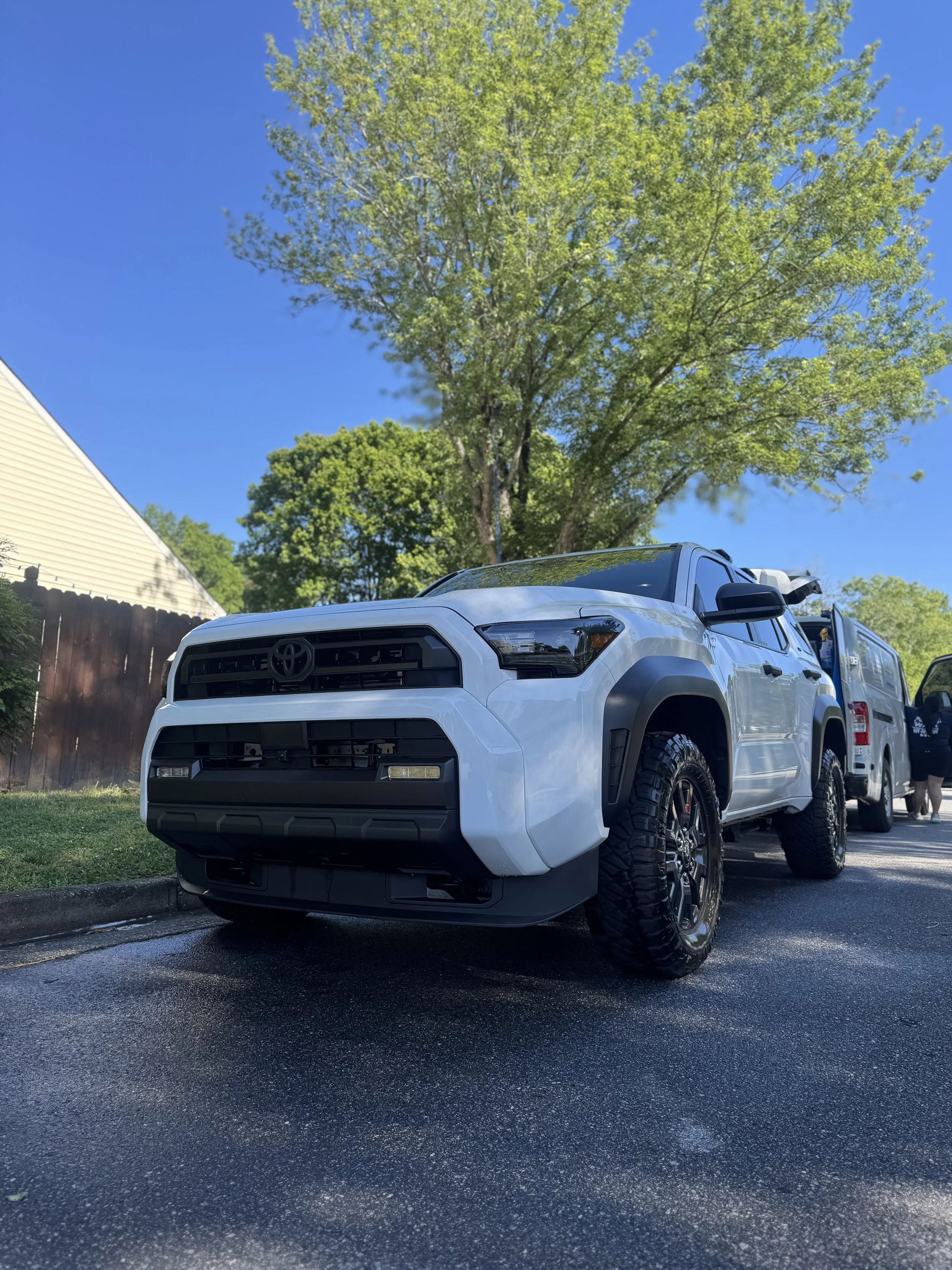 A white Toyota Tacoma pickup truck parked on a wet asphalt surface in a residential area. There are trees with green leaves and a clear blue sky in the background.