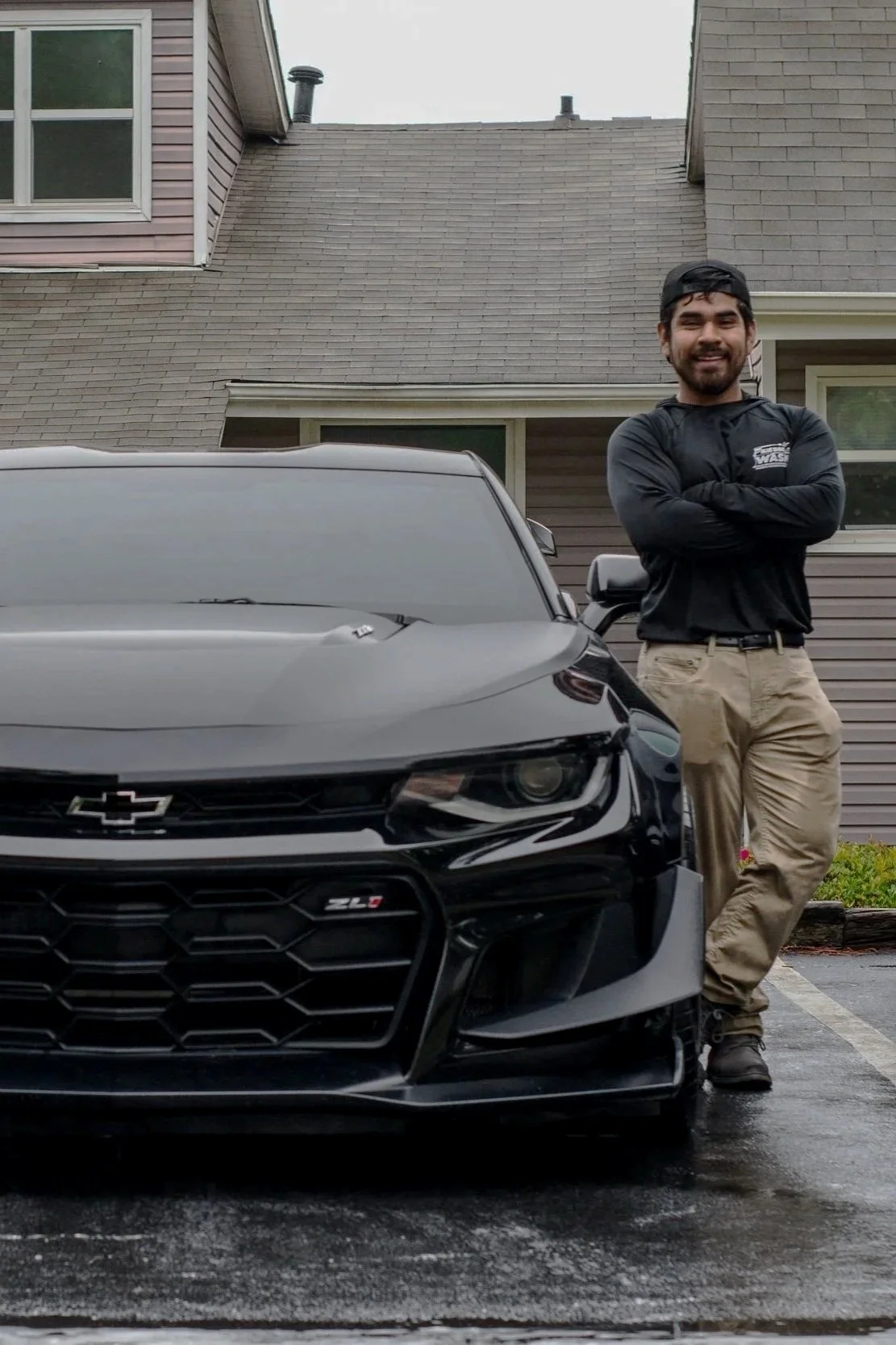 A man with a beard, wearing a black jacket and beige pants, standing cross-armed next to a black Chevrolet Camaro ZL1 parked in front of a residential building with brown siding. The man is smiling.