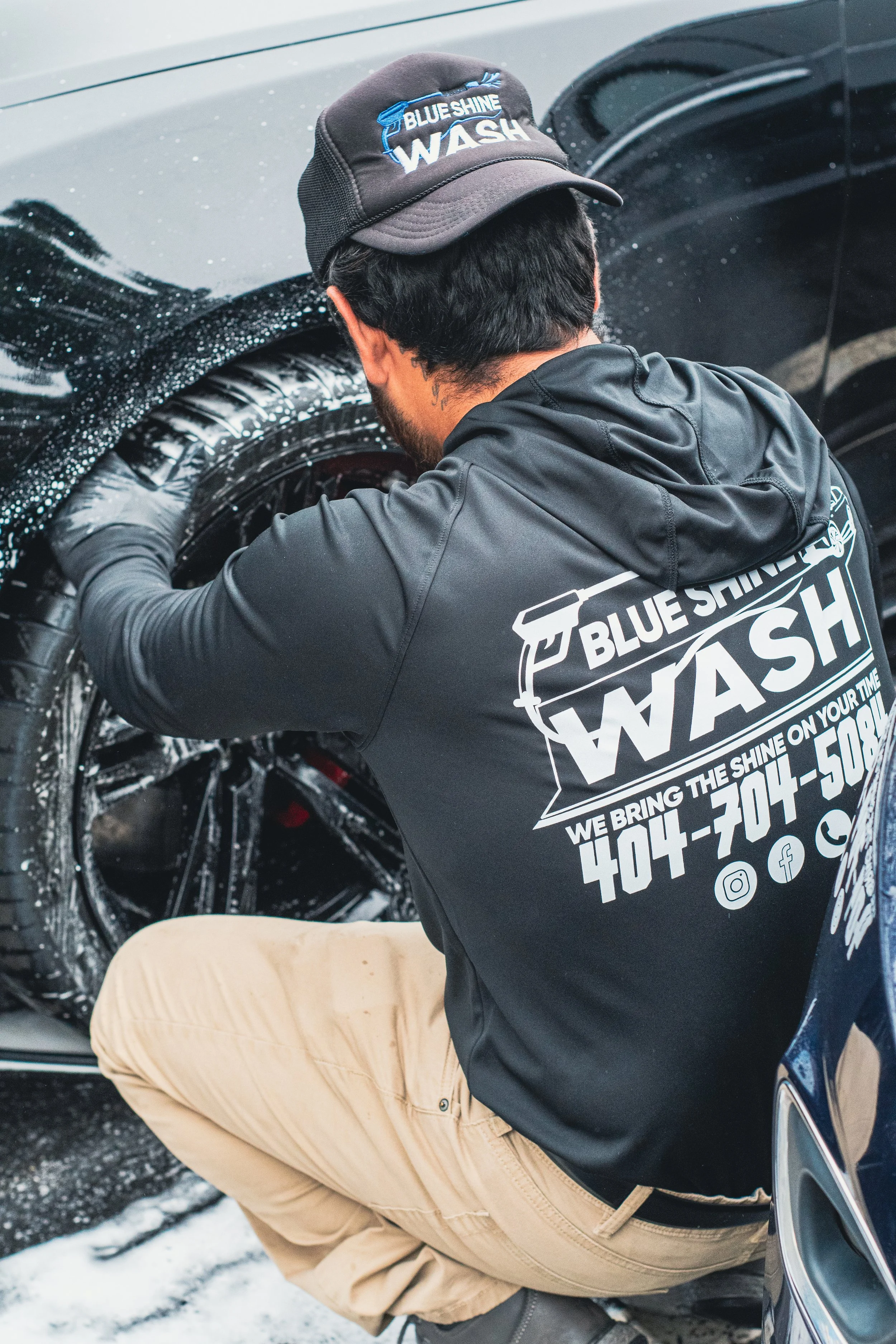 Person wearing a black jacket and beige pants washing a car with foam, wearing a black 'Blue Shine Wash' cap, kneeling on the ground.