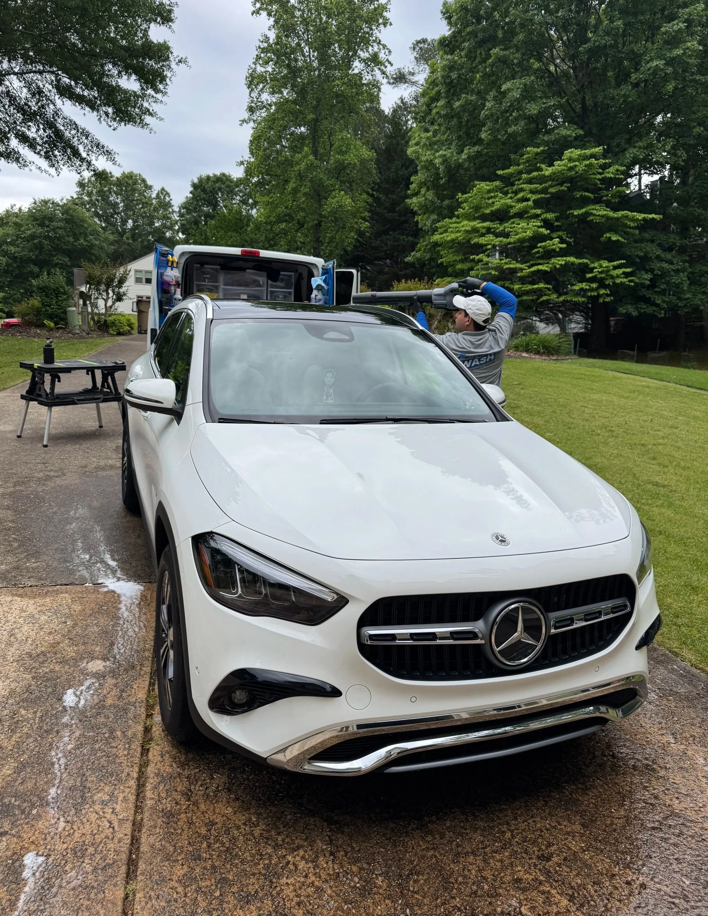A man cleaning the roof of a white Mercedes-Benz car with a leaf blower in a suburban driveway during daytime. There is a work table and a van with open doors behind him, and trees and a house are visible in the background.