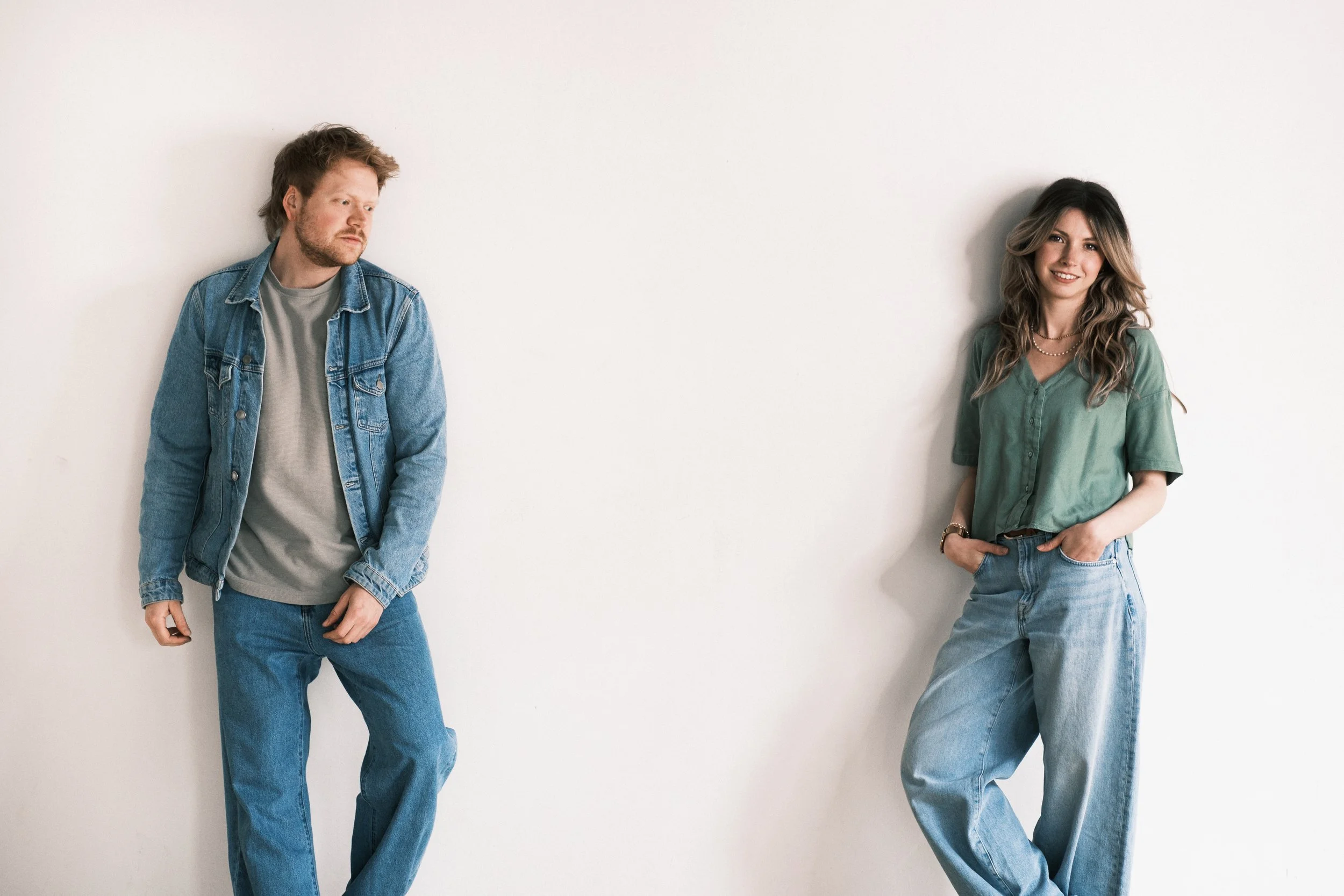 Ein Mann und eine Frau lehnen an einer weißen Wand. Der Mann trägt eine Denim-Jacke, das Mädchen trägt eine grüne Bluse und Jeans. Beide wirken entspannt.