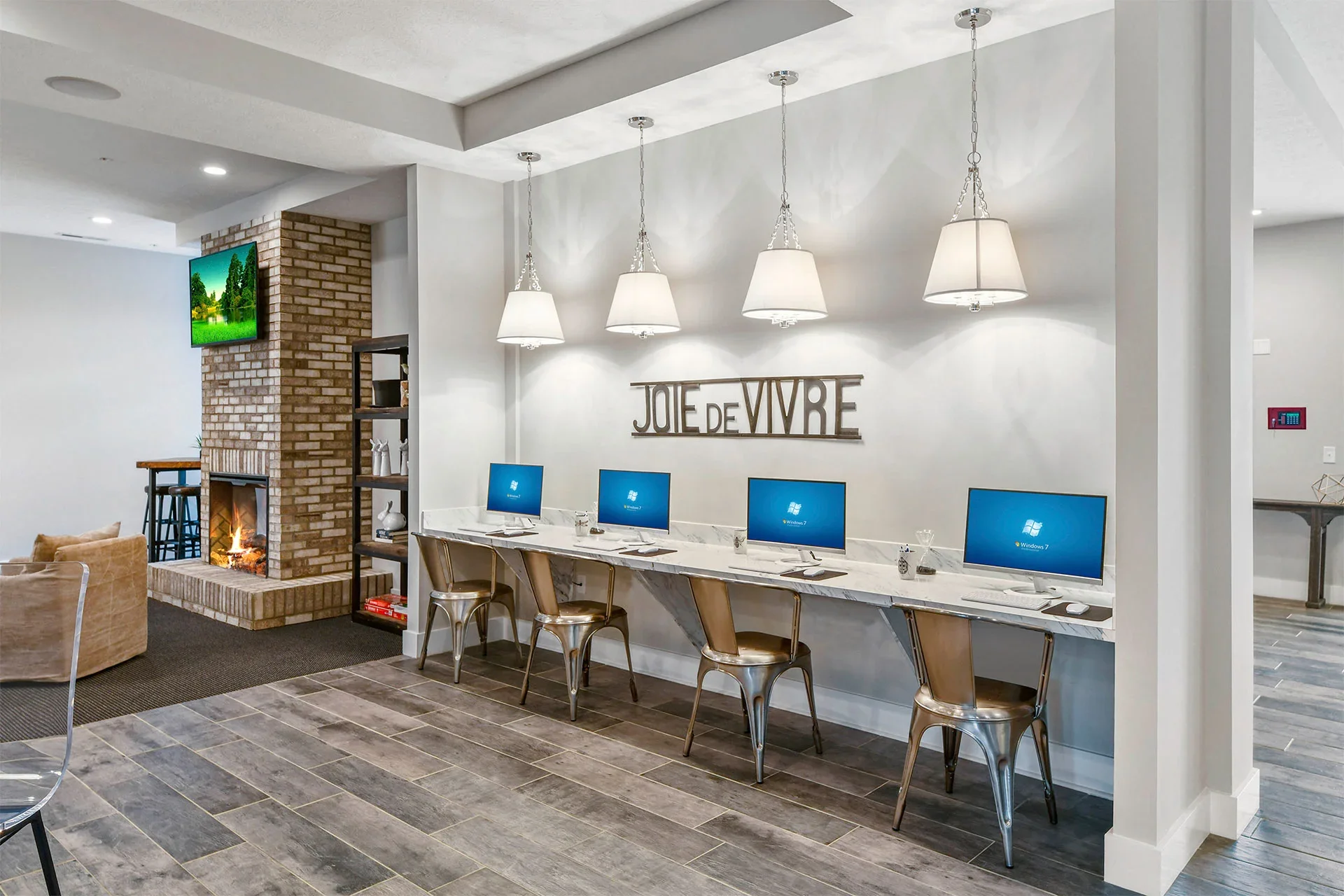 Modern shared workspace with four Apple computers on a marble counter, silver chairs, white pendant lights, and a decorative sign on the wall that reads 'Joie de Vivre'. Adjacent is a brick fireplace with a TV mounted above and a sitting area with a 