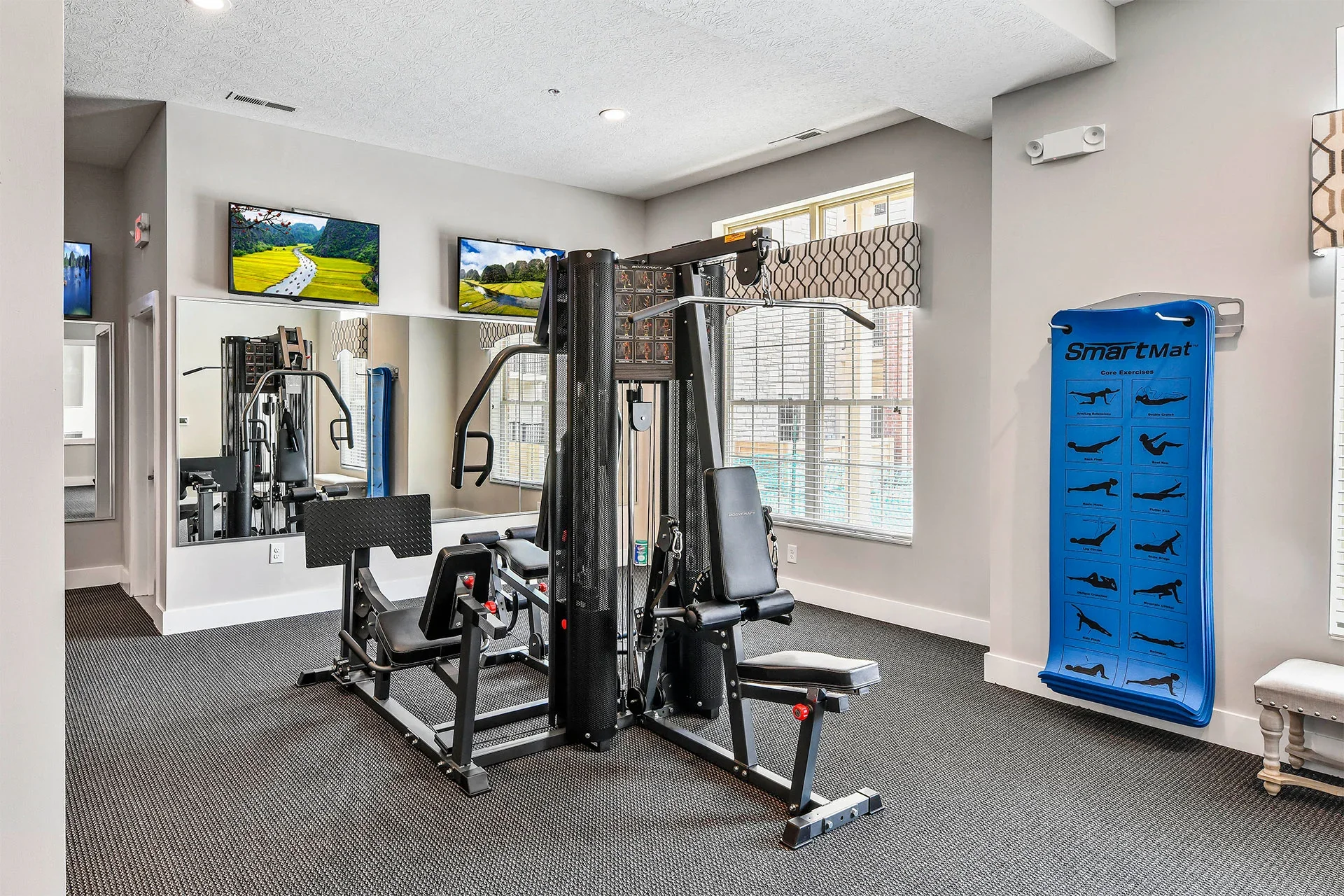 Indoor gym with workout equipment, wall-mounted televisions, and a blue exercise diagram chart on the wall.