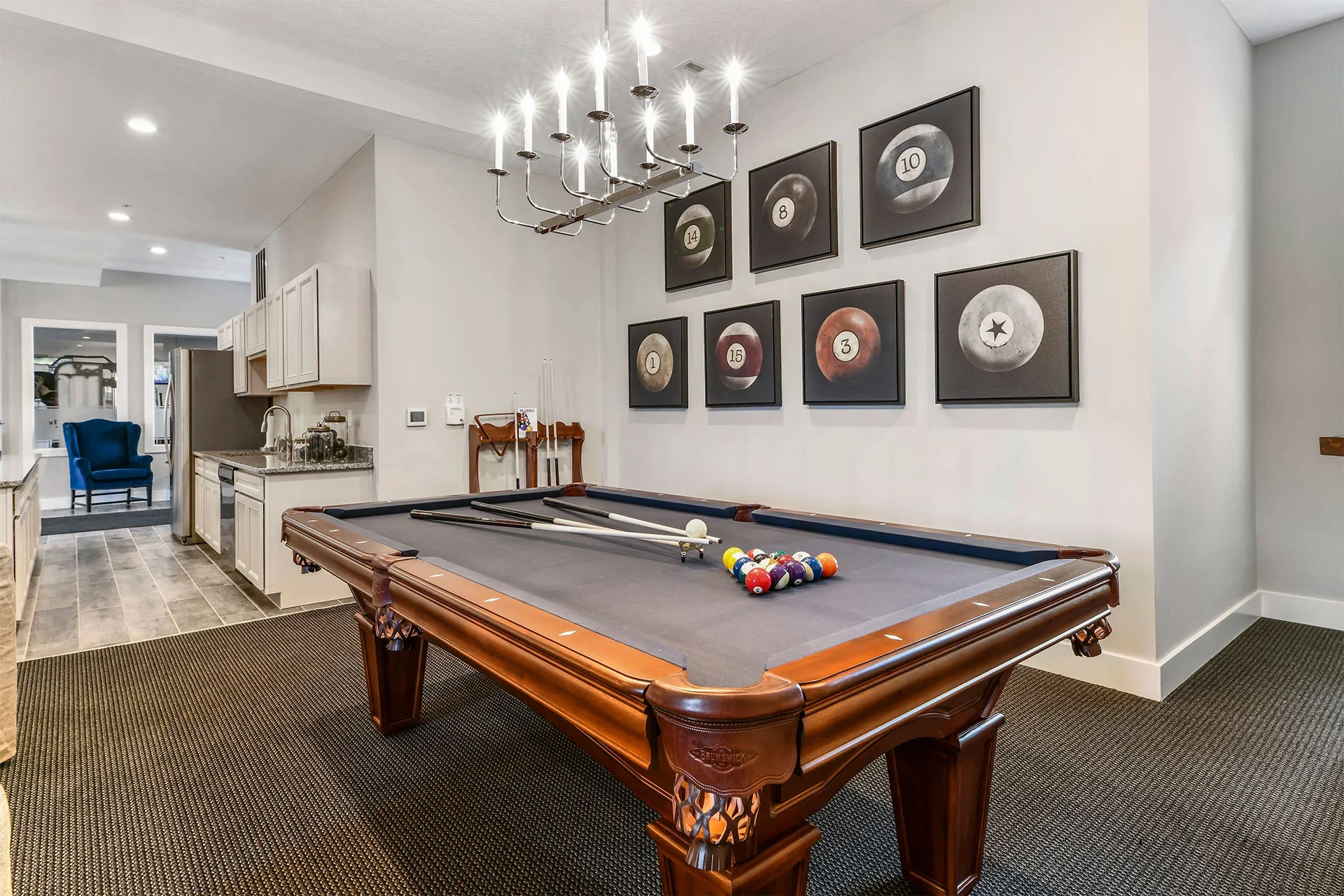 A pool table in a room with framed billiard ball pictures on the wall, a chandelier overhead, and a kitchenette in the background.
