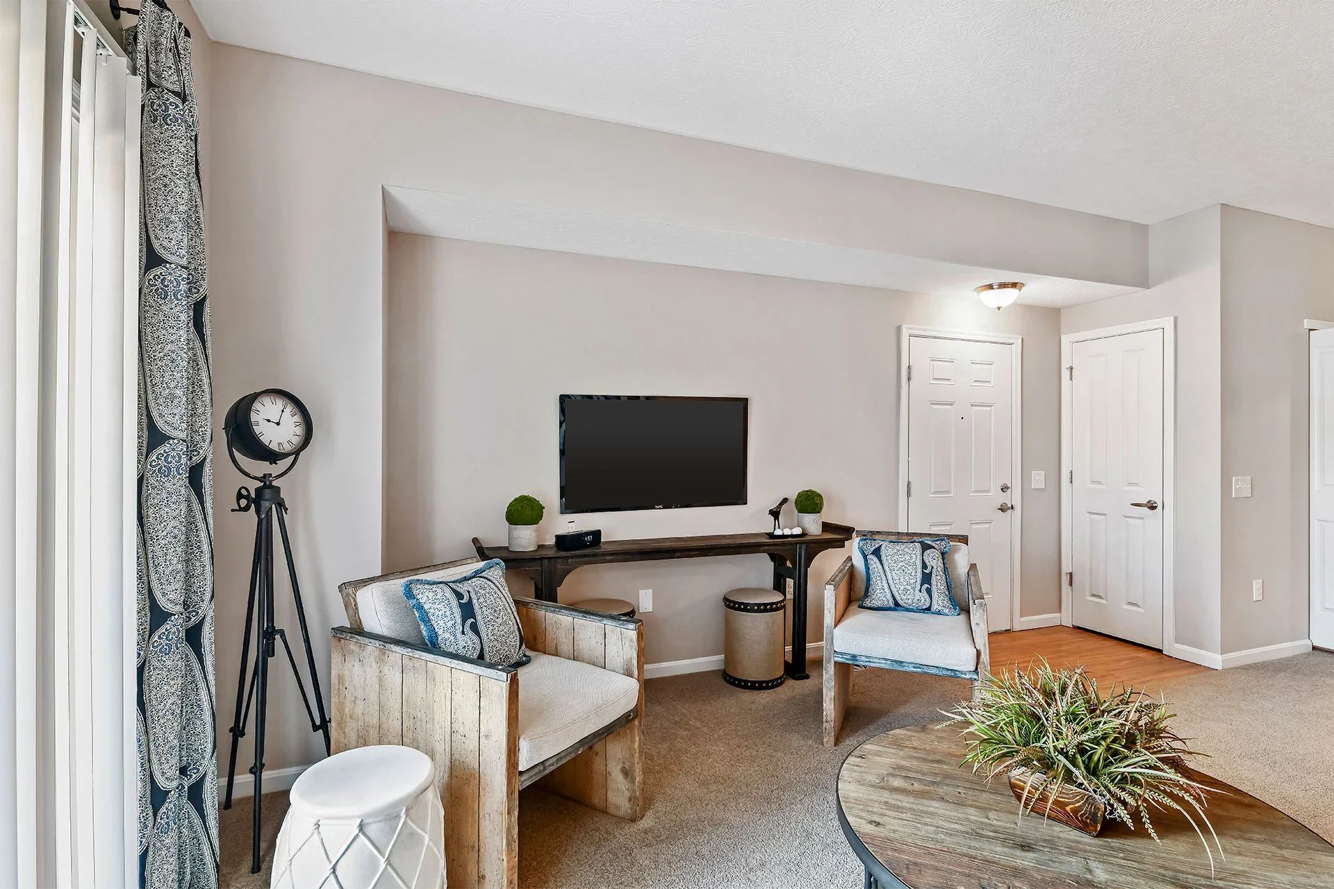 A living room with a flat-screen TV mounted on a beige wall, two armchairs with blue and white cushions, a wooden table with decorative plants, and a window with patterned curtains.