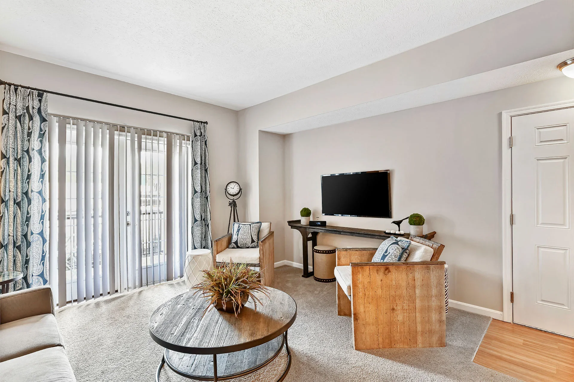 Living room with sliding glass door, curtains, chairs, a table with a plant, a wall-mounted TV, and decorative items.
