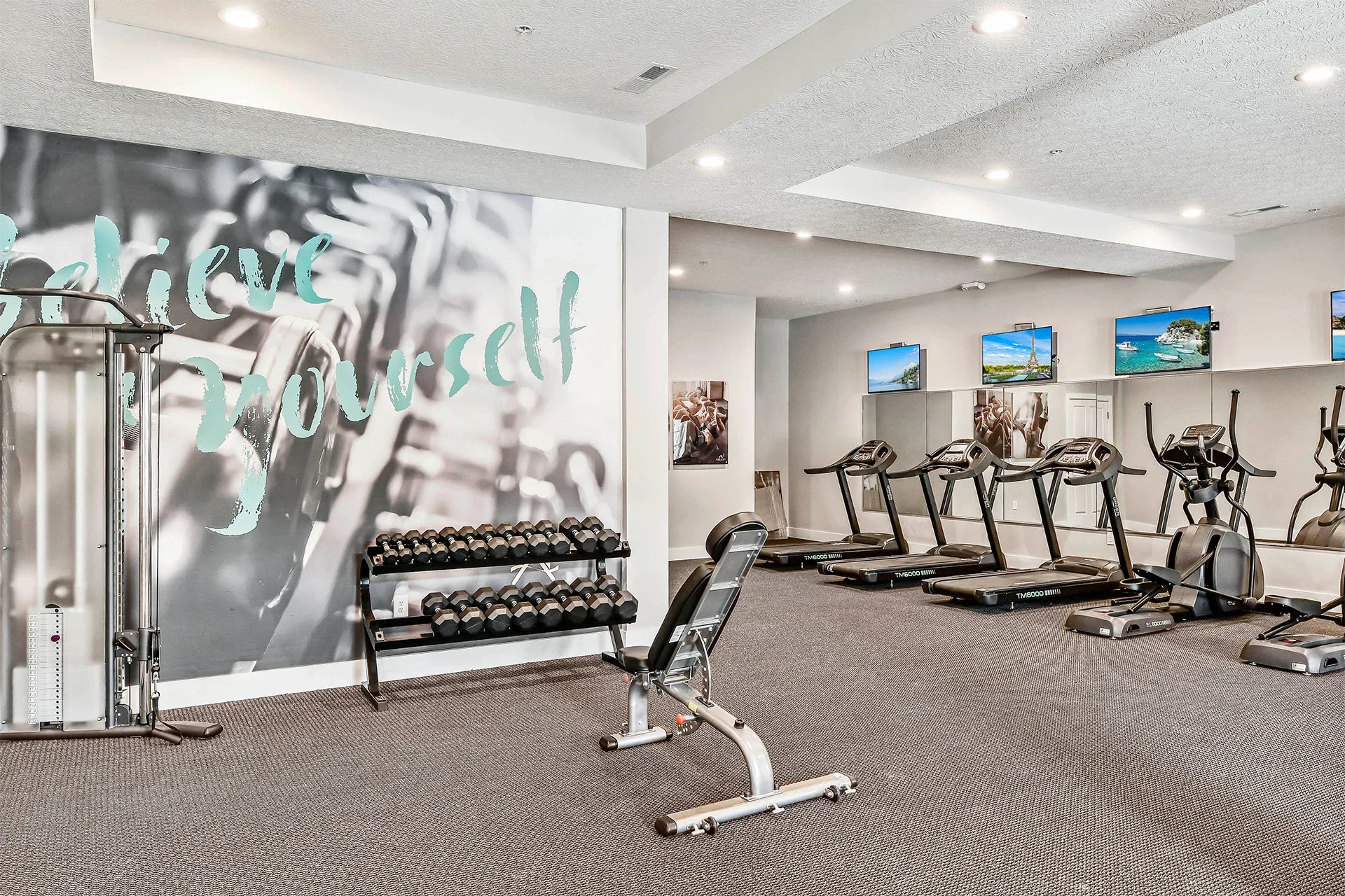 A modern gym with treadmills, ellipticals, a rack of dumbbells, and wall-mounted televisions showing scenic views and landmarks.